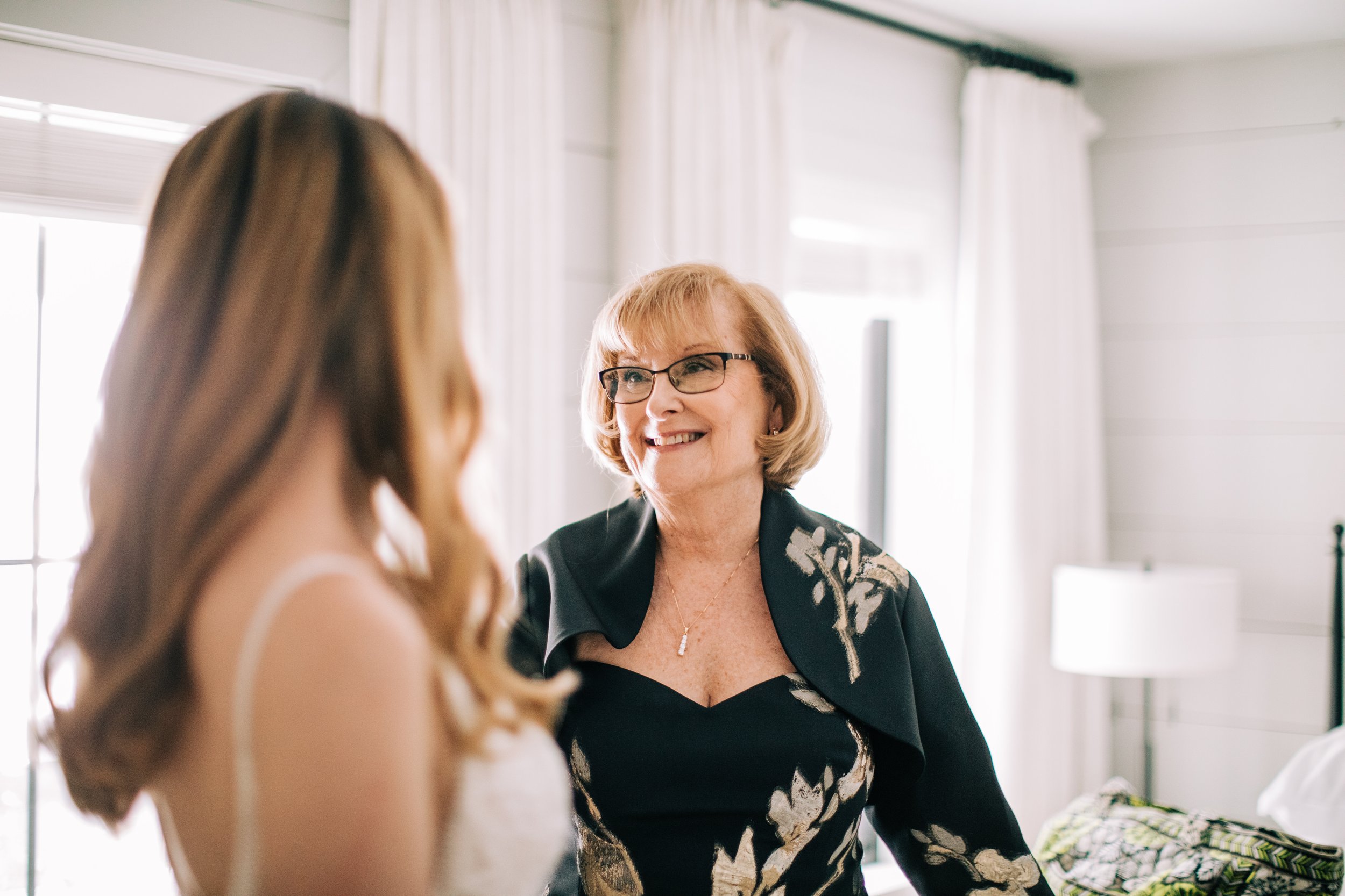 A smiling older woman with glasses, short blonde hair, and a floral black dress talking to a woman with long brown hair in a white dress in a bright room with white curtains and a bed.