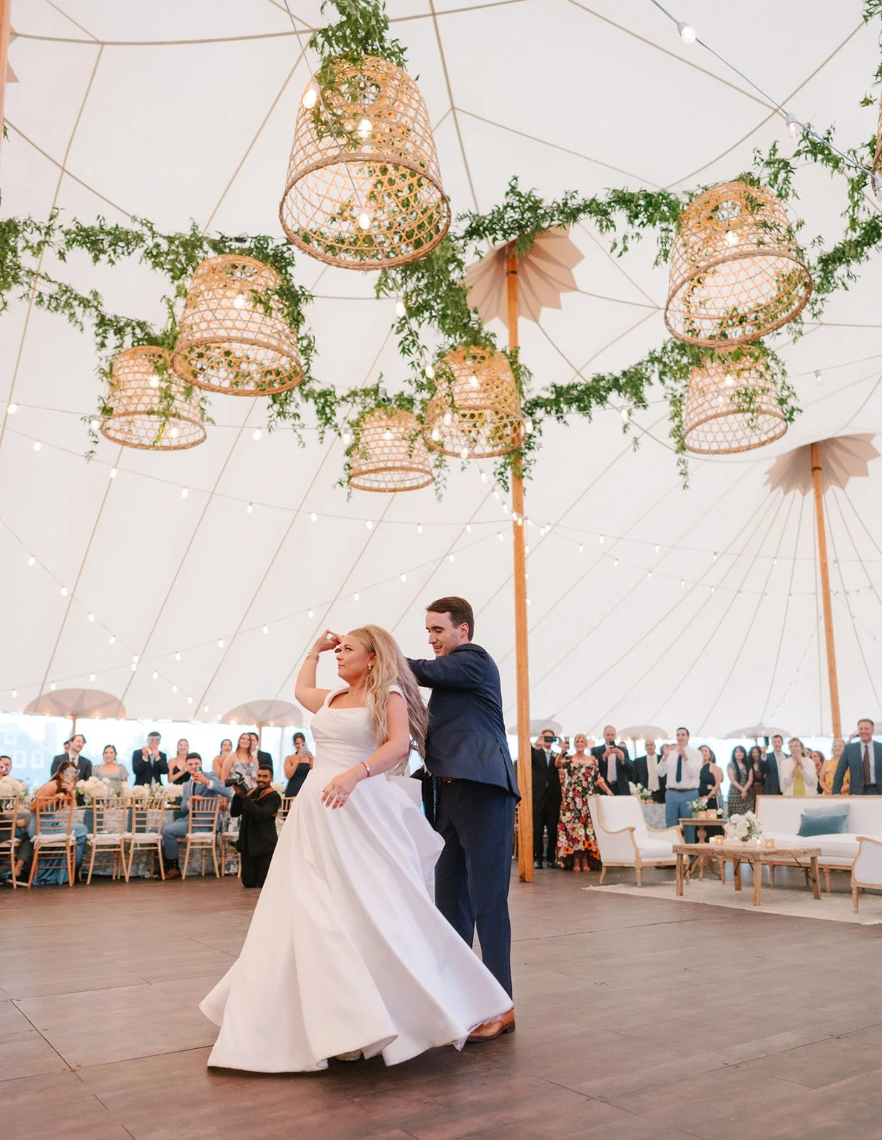 A bride and groom are dancing at their wedding reception inside a large tent decorated with hanging wicker lanterns and greenery. Guests are watching and taking photos in the background.