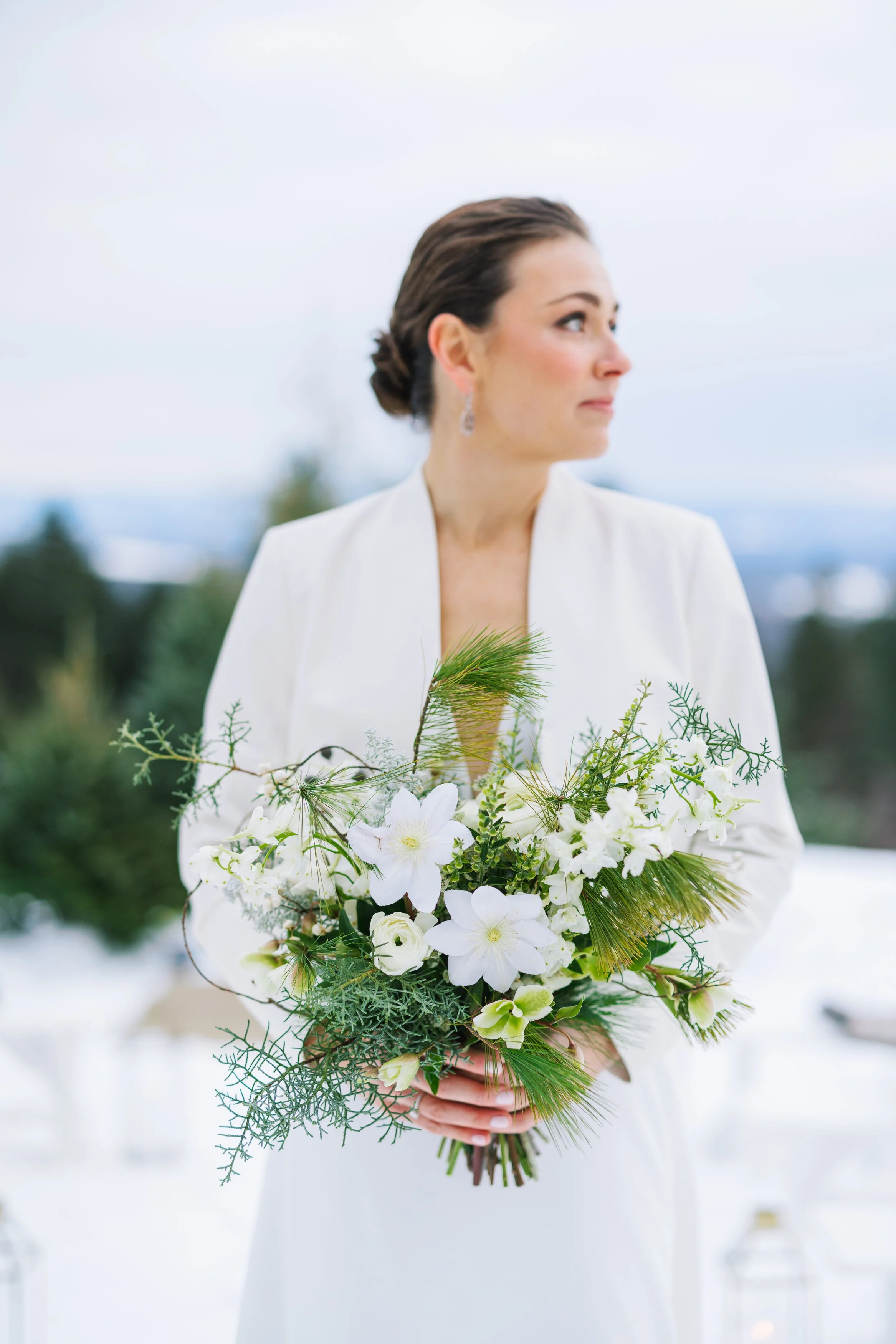 A woman in a white dress holding a bouquet of white flowers and greenery outdoors on a cloudy day.