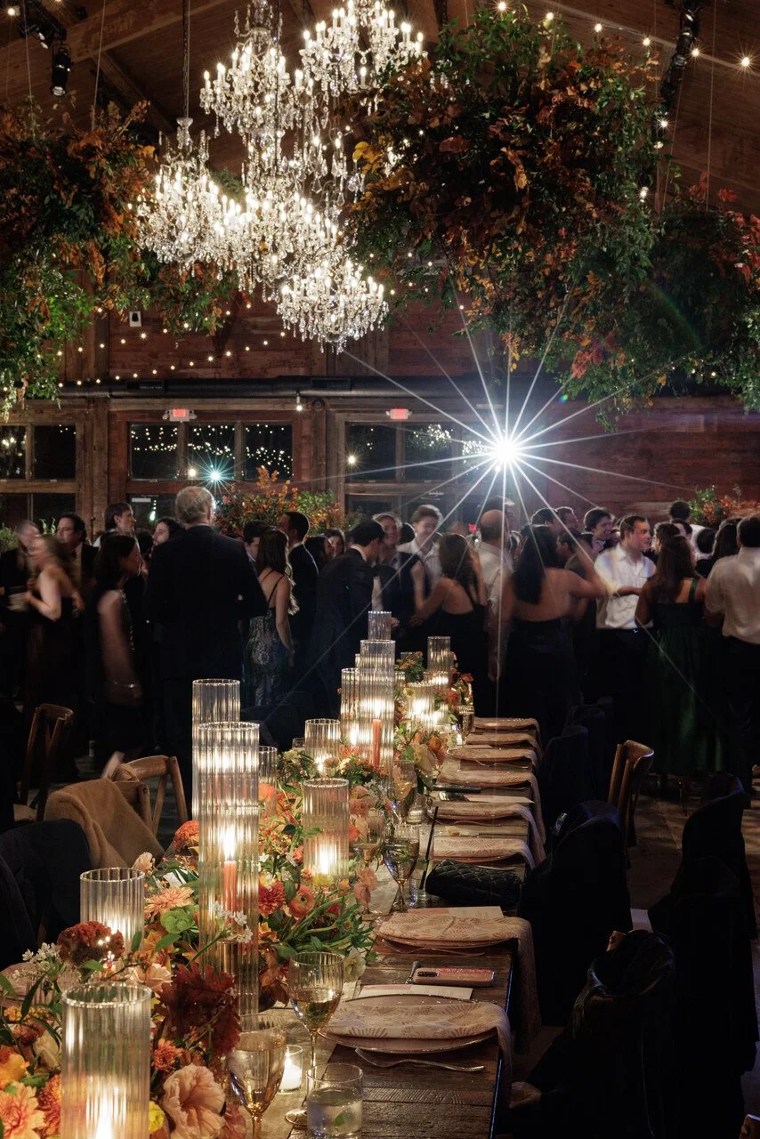 An elegant indoor wedding reception with a long dining table decorated with floral arrangements and tall candle holders. Guests are dancing under hanging chandeliers and string lights, with a rustic wooden interior and large windows in the background