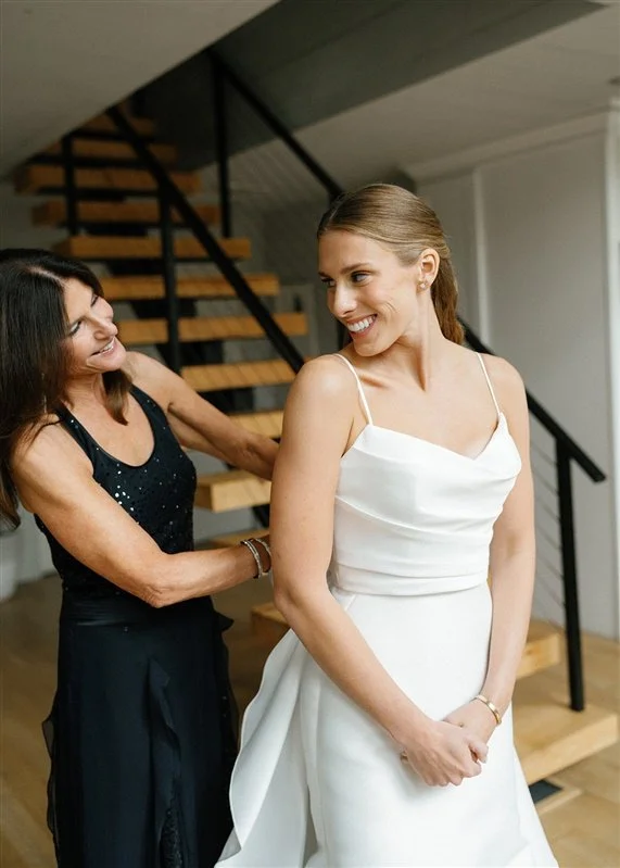 A woman in a white dress is smiling as another woman in a black dress helps her adjust her gown, standing near a staircase indoors.