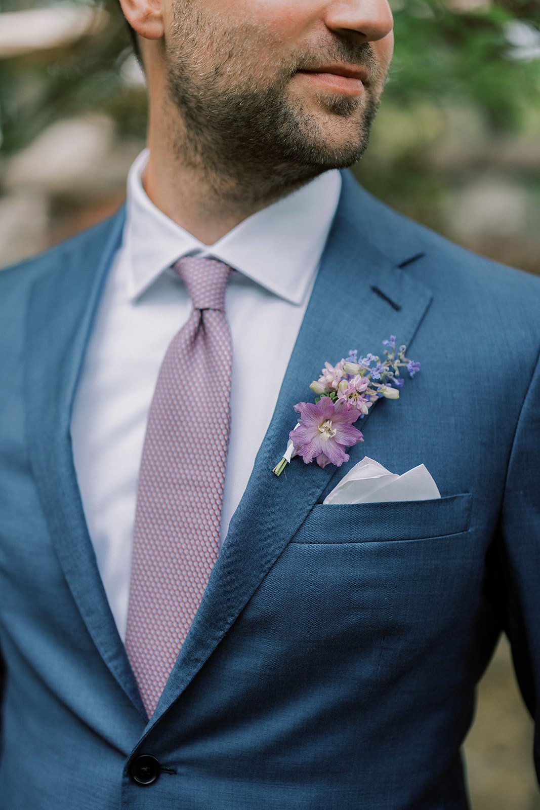 Close-up of a man in a blue suit with a white shirt and a pink tie, with a pink and purple boutonniere on his lapel and a white pocket square in his breast pocket.