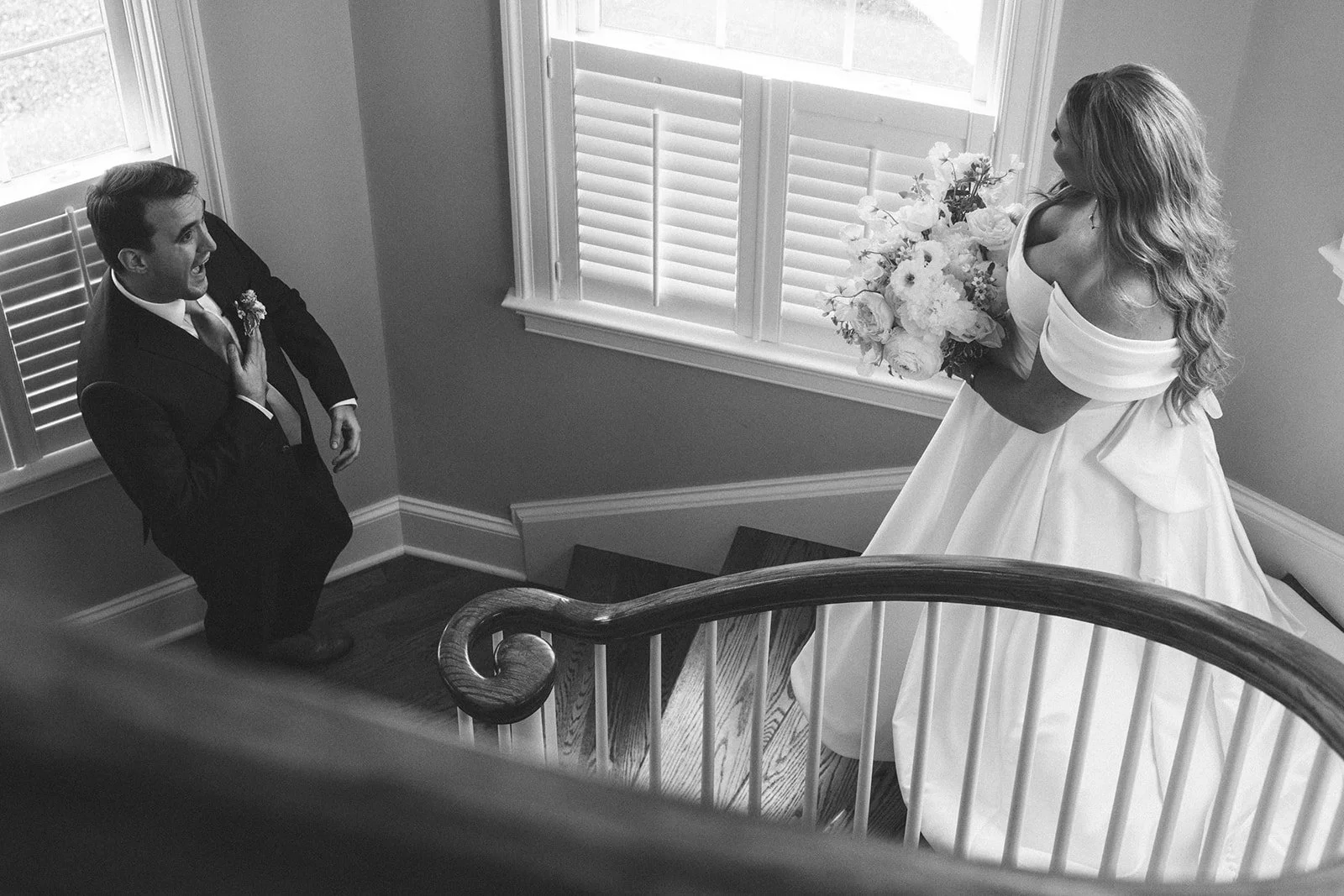 A woman in a white wedding dress holding a bouquet of flowers on a staircase, looking at a man in a suit kneeling with one hand on her chest, inside a house with windows and shutters.