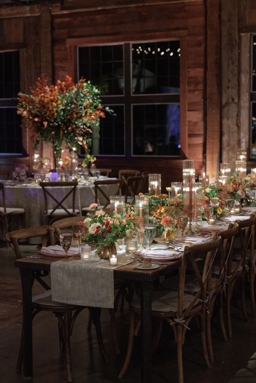 A beautifully decorated indoor wedding reception table with flowers, candles, and elegant table settings in a rustic wooden venue at night.