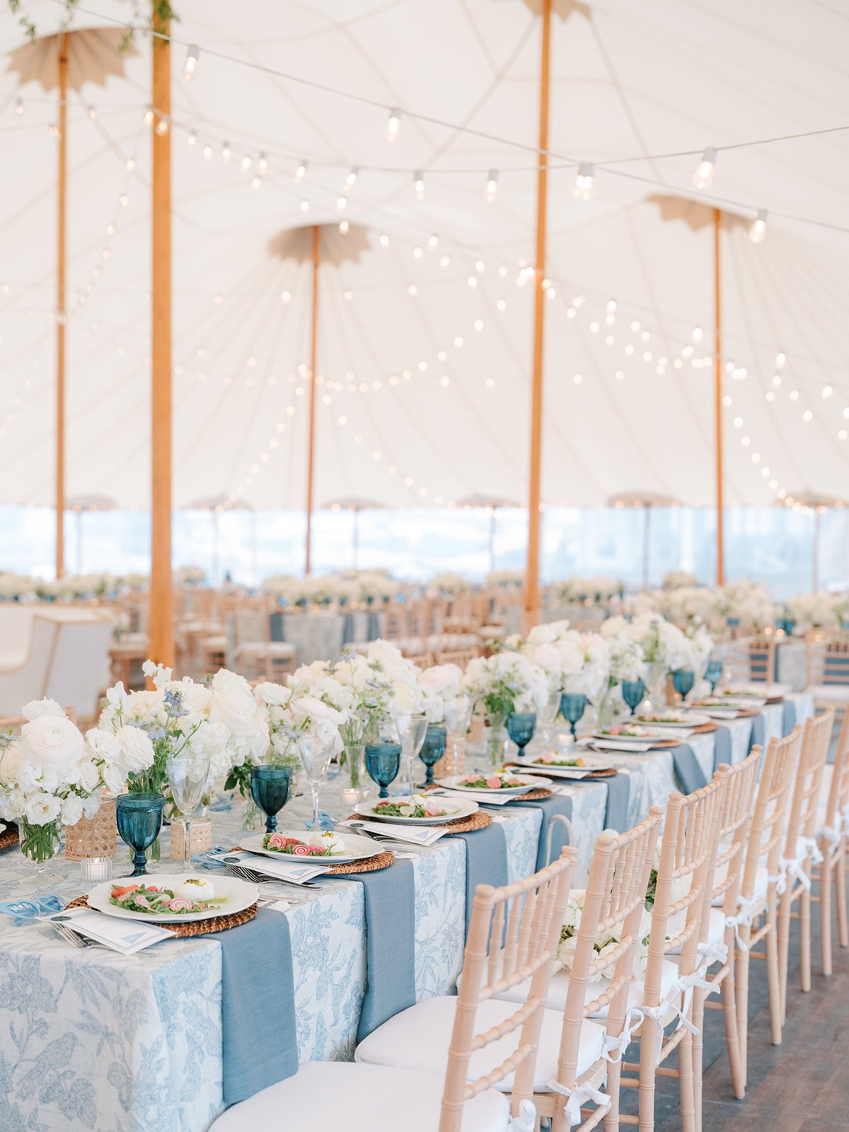 Elegant outdoor dining setup under a tent with string lights, decorated with white floral arrangements and tableware with blue accent glasses.