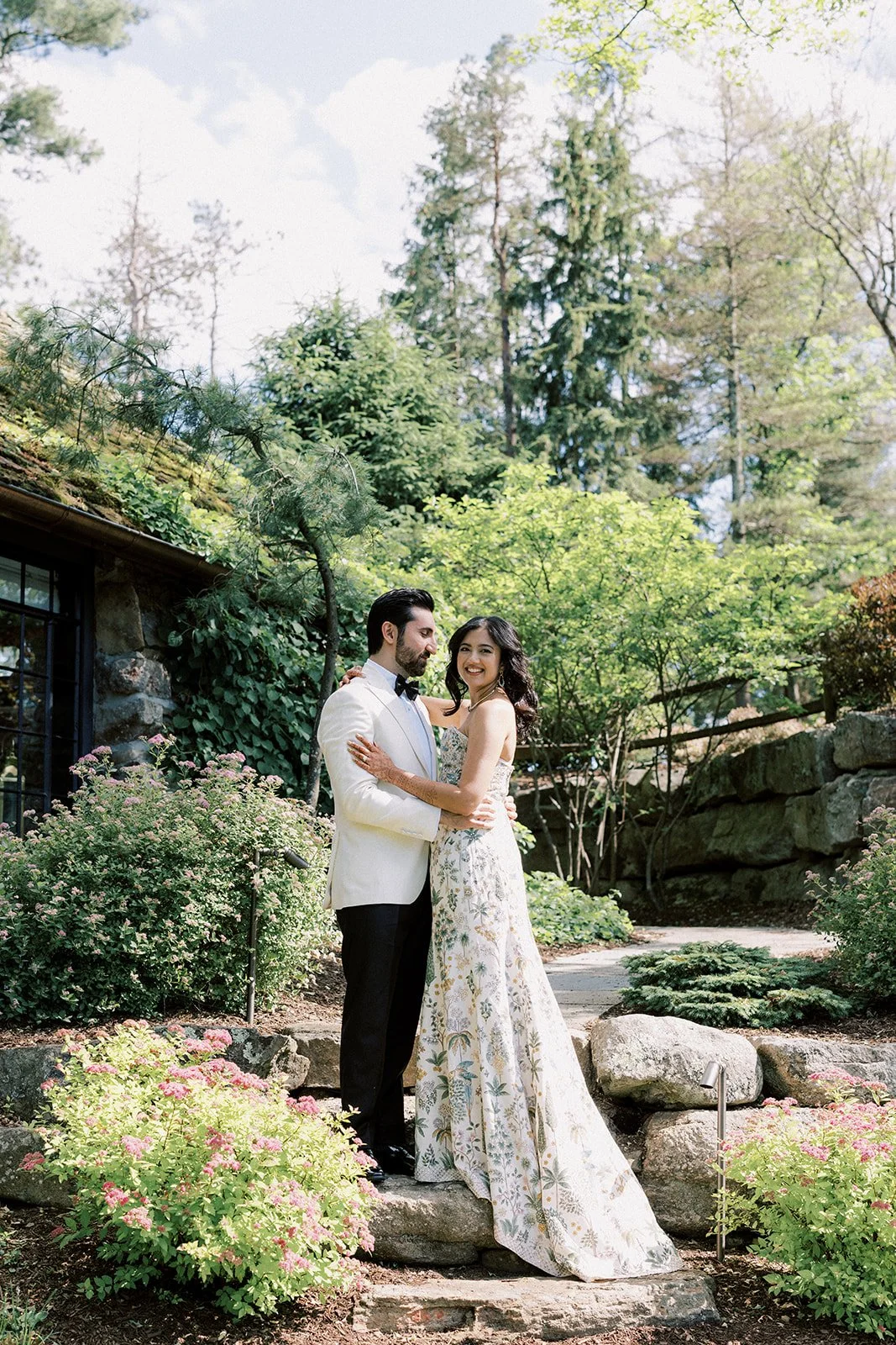 A couple dressed in formal wedding attire standing on stone steps in a lush garden, embracing and smiling at each other surrounded by green trees, bushes, and flowers.