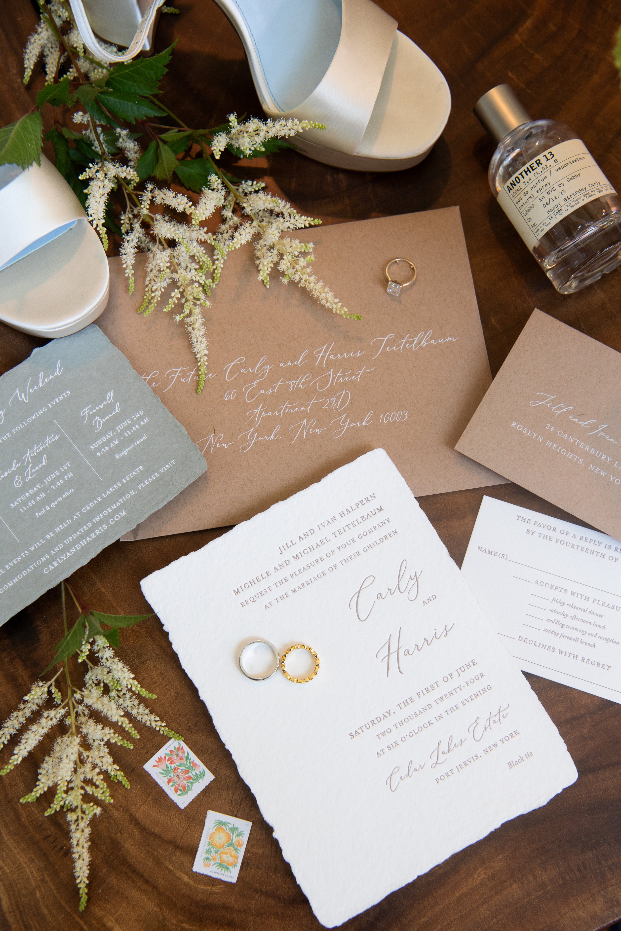 Wedding invitation, rings, a rose gold ring and a silver ring, a perfume bottle, white high-heeled shoes, greenery, and decorative stickers on a wooden surface.