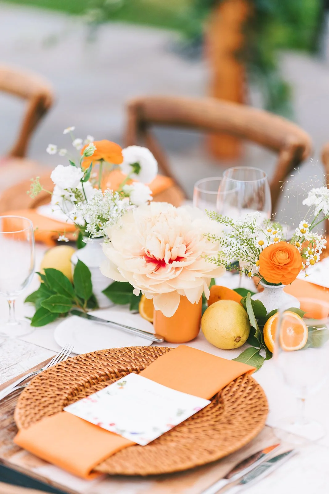 Table setting with orange napkins, floral centerpieces with white, orange, and pink flowers, lemon, and orange slices at an outdoor event.