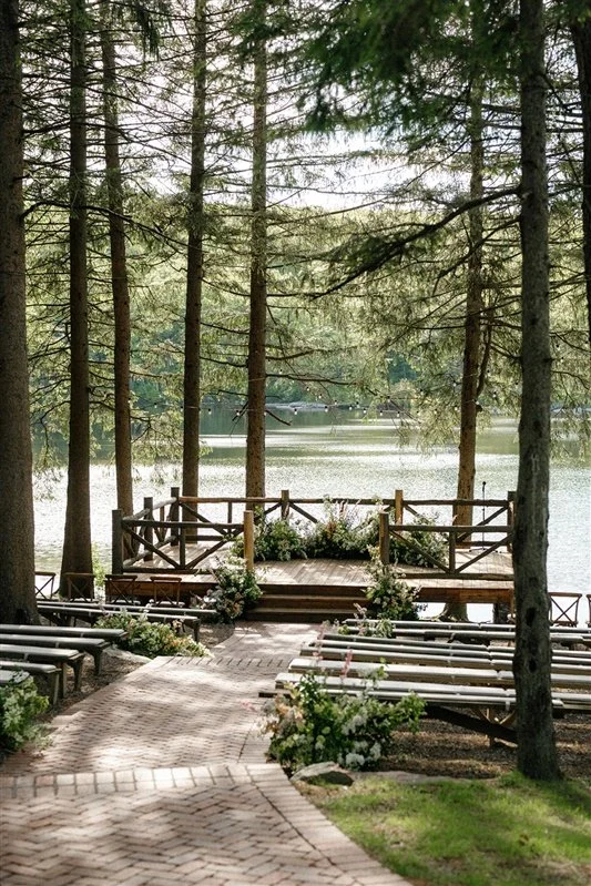 A lakeside outdoor venue with a wooden stage, surrounded by pine trees and floral arrangements, overlooking a calm lake.
