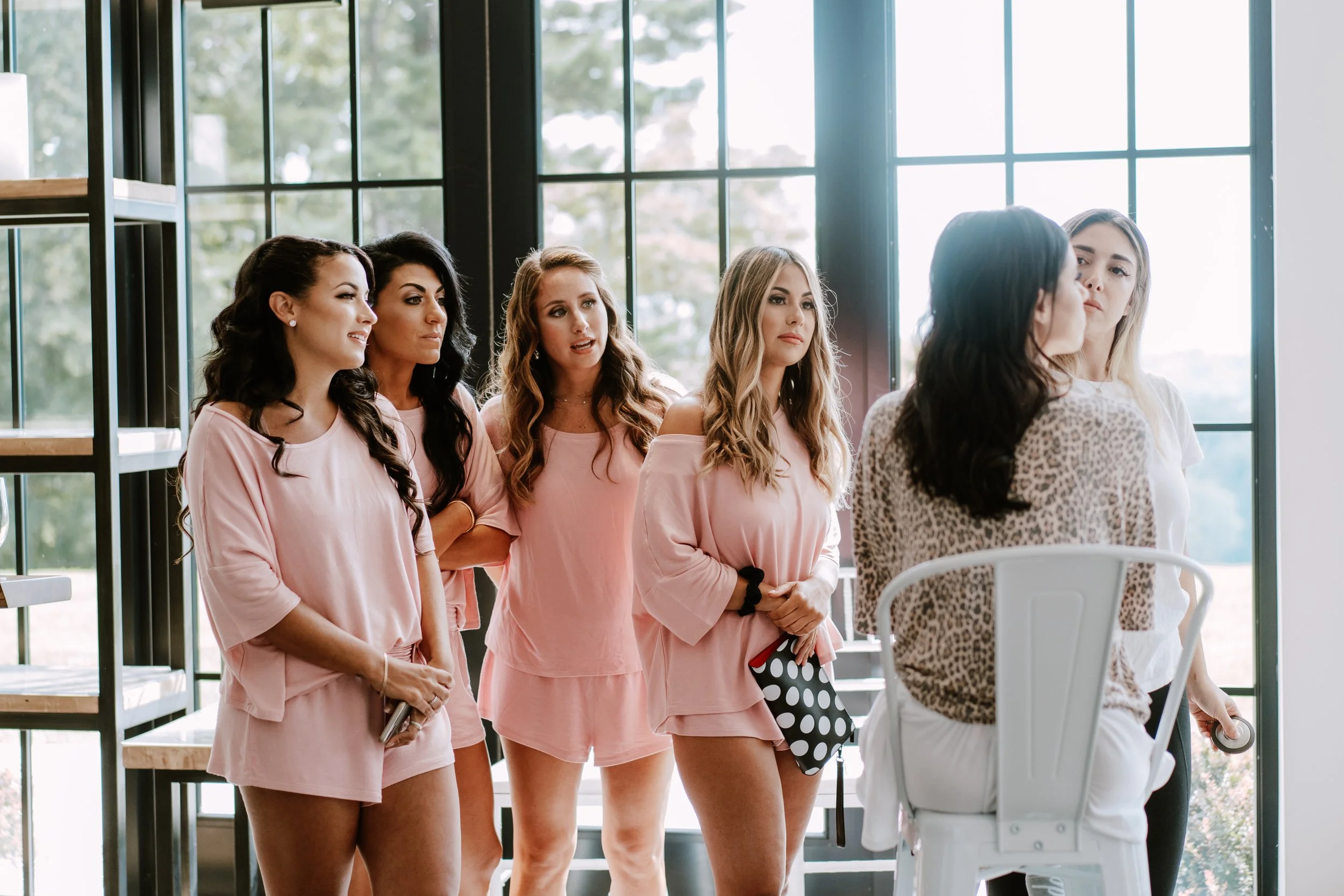 Group of women standing in line inside a bright room with large windows, dressed in pink casual loungewear, engaged in conversation with a woman seated facing them.