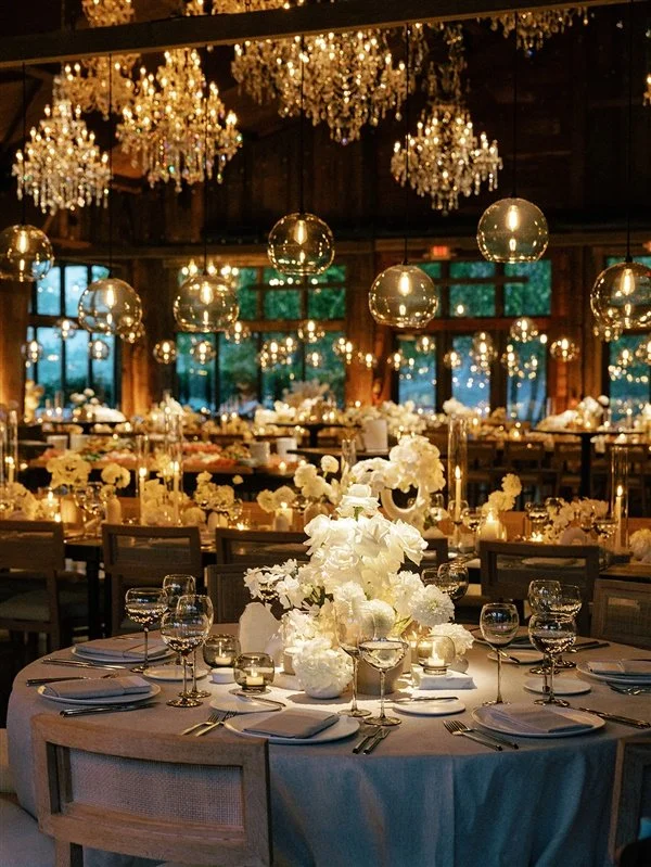 Elegant banquet hall decorated with white floral centerpieces, gold candle holders, and hanging chandeliers, with round tables set with white napkins, glassware, and silverware.