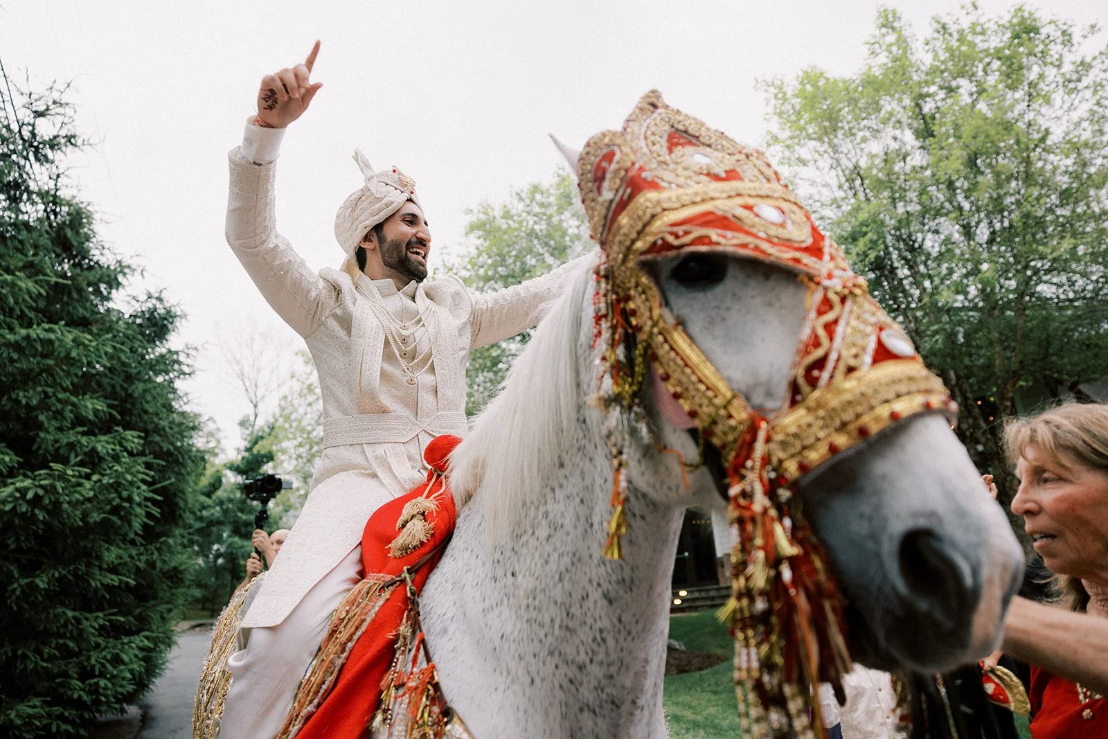 A man dressed in traditional Indian wedding attire riding a white horse decorated with colorful and ornate accessories, waving and smiling during a wedding celebration.