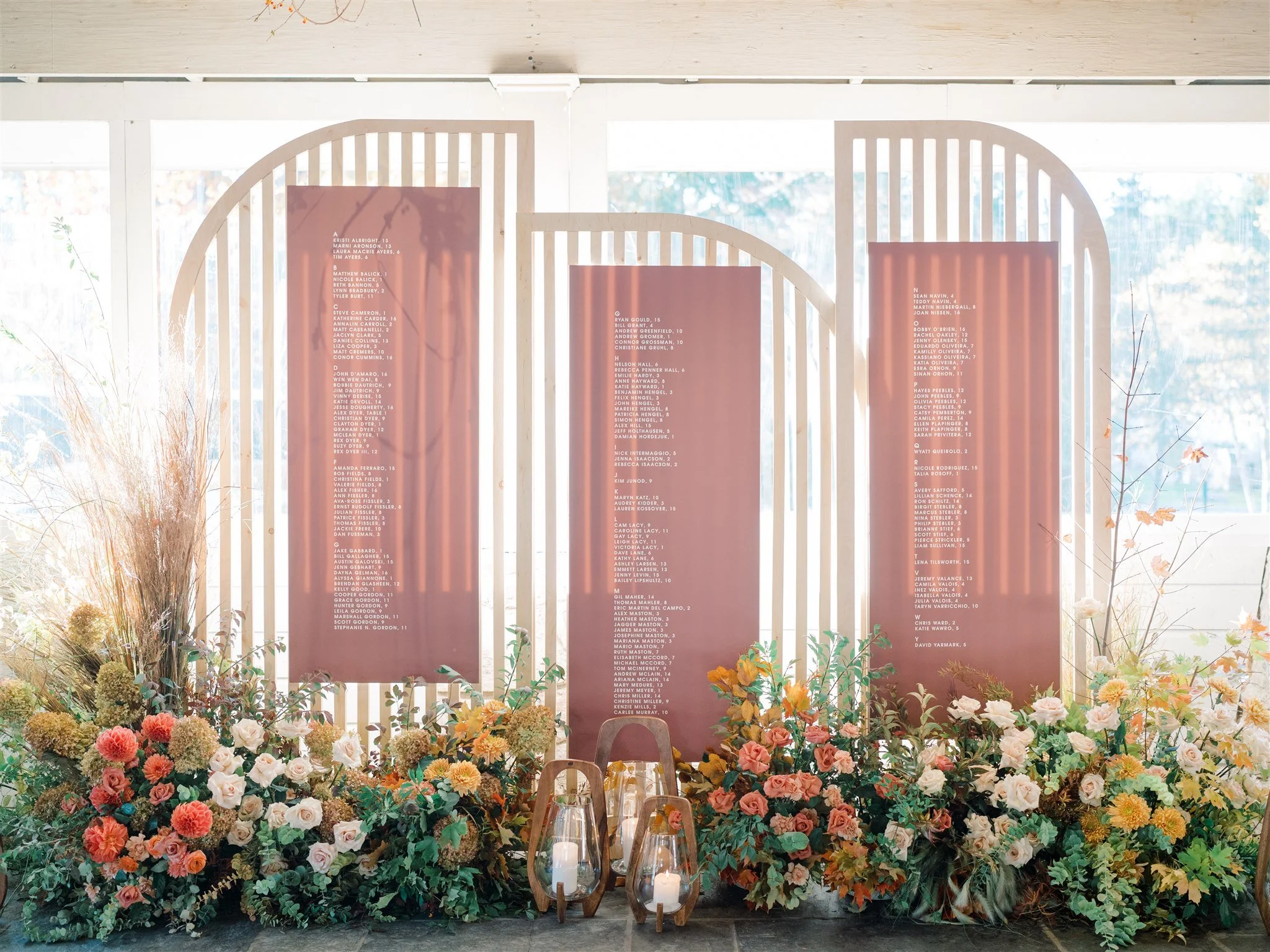 A display of three large, vertical pink boards with white text arranged in front of a light-colored wooden backdrop with vertical slats. The boards contain lists of names. At the base of the display, there are numerous colorful flowers and green foli