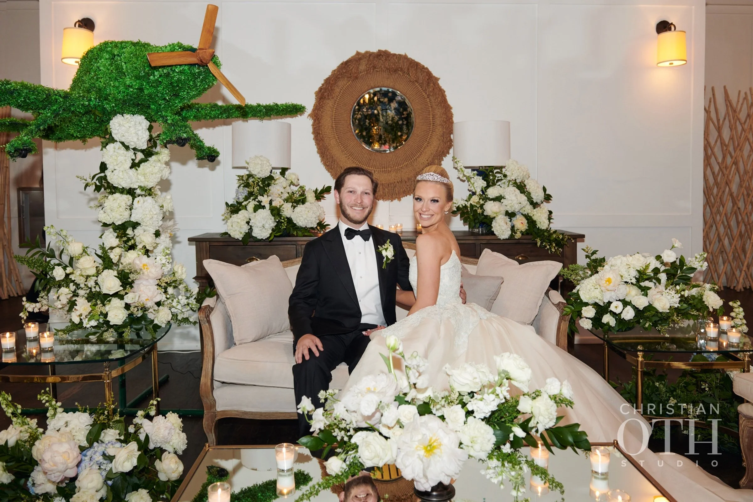 A newly married couple sitting on a cream-colored sofa, surrounded by white floral arrangements and candles, in a decorated indoor setting with a green helicopter prop on the wall behind them.