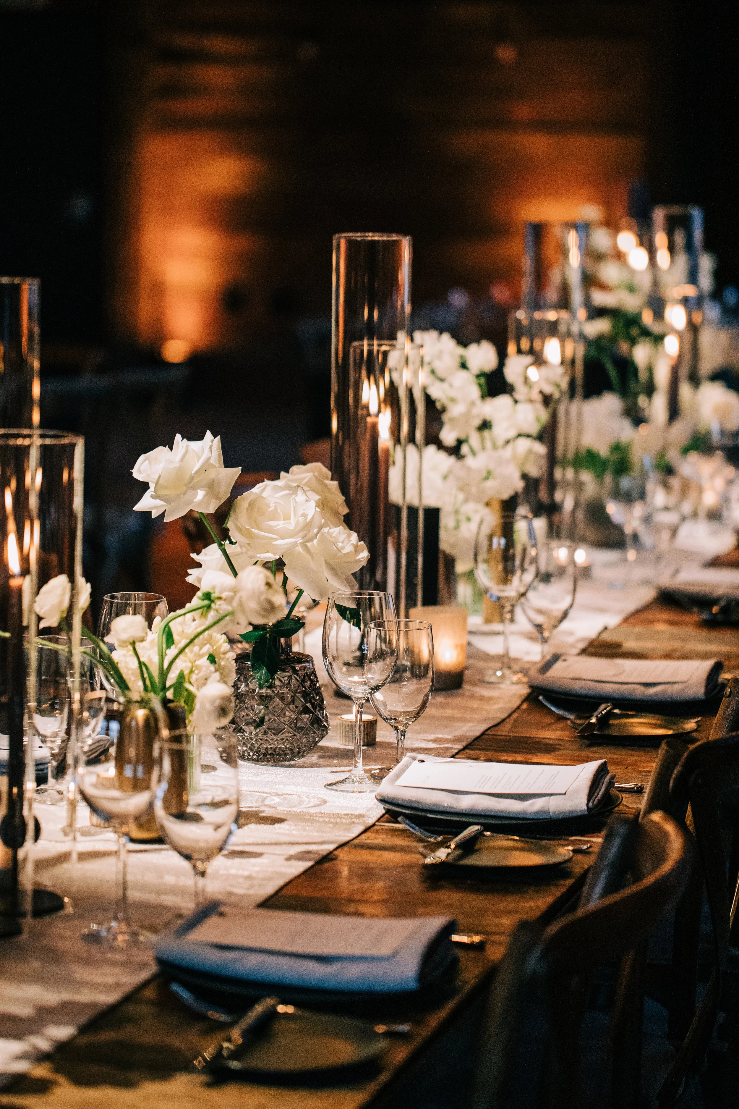 Elegant dining table set for a formal event with white flowers, candles, glassware, and napkins in a dimly lit room.