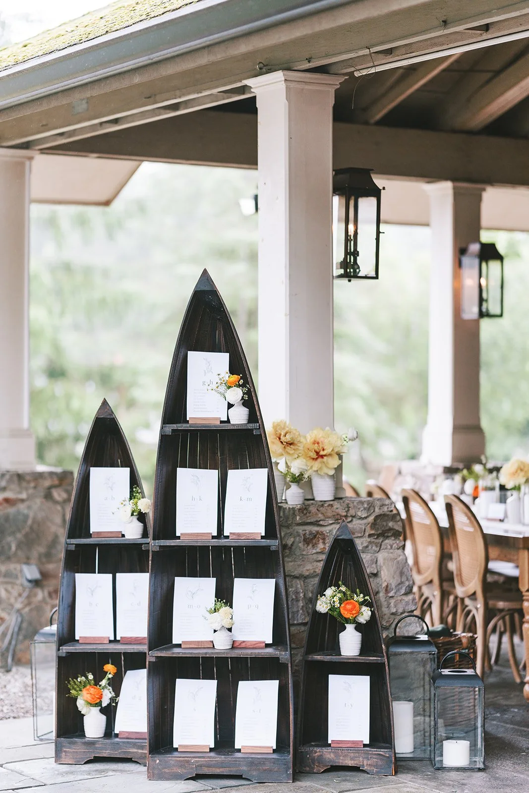 Table seating chart display with dark wooden boat-shaped shelves decorated with small flower arrangements, set against a stone wall and patio with tables and chairs in the background.