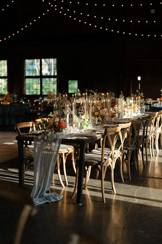 Wedding reception table decorated with floral arrangements and candles, under string lights in a rustic barn setting.