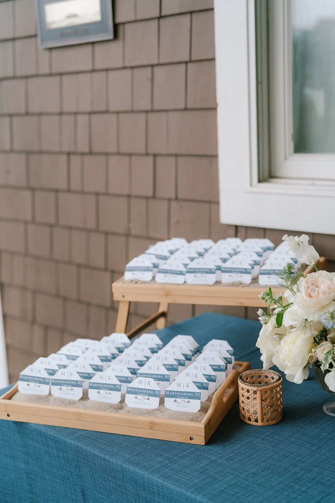 Table with a teal tablecloth displaying name tags, a bouquet of white and pink flowers, and a small decorative basket near a window with brown shingle siding outside.