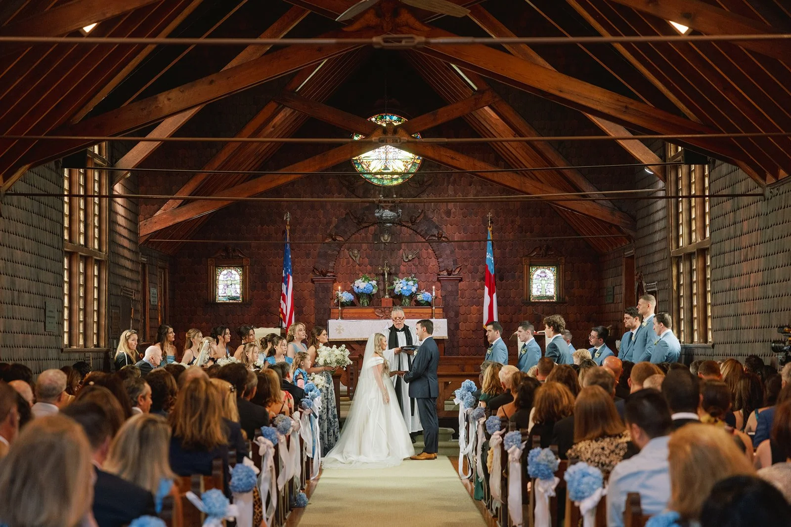 A wedding ceremony inside a church with wooden interior, with bride and groom standing at the altar. The officiant reads from a book, surrounded by bridesmaids and groomsmen. Guests are seated in rows, decorated with blue flowers. Two American flags 