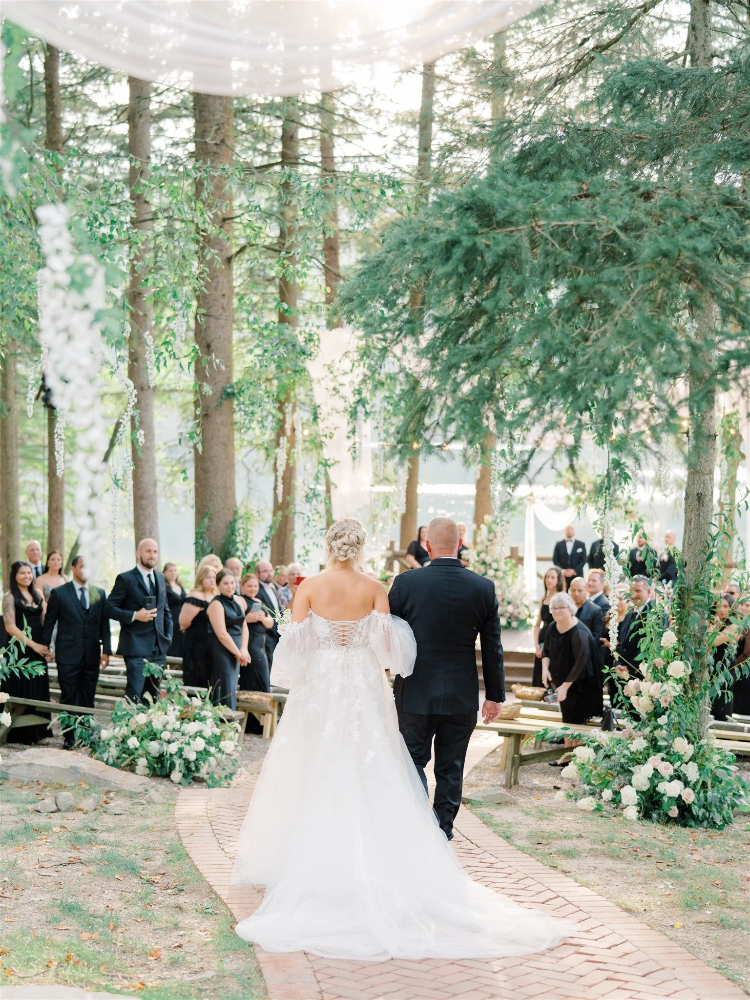 Bride and groom walking down the aisle at an outdoor wedding ceremony in a wooded area. The bride is in a white gown with off-the-shoulder sleeves, and the groom is in a black suit. Guests are seated on both sides, with floral decorations and greener