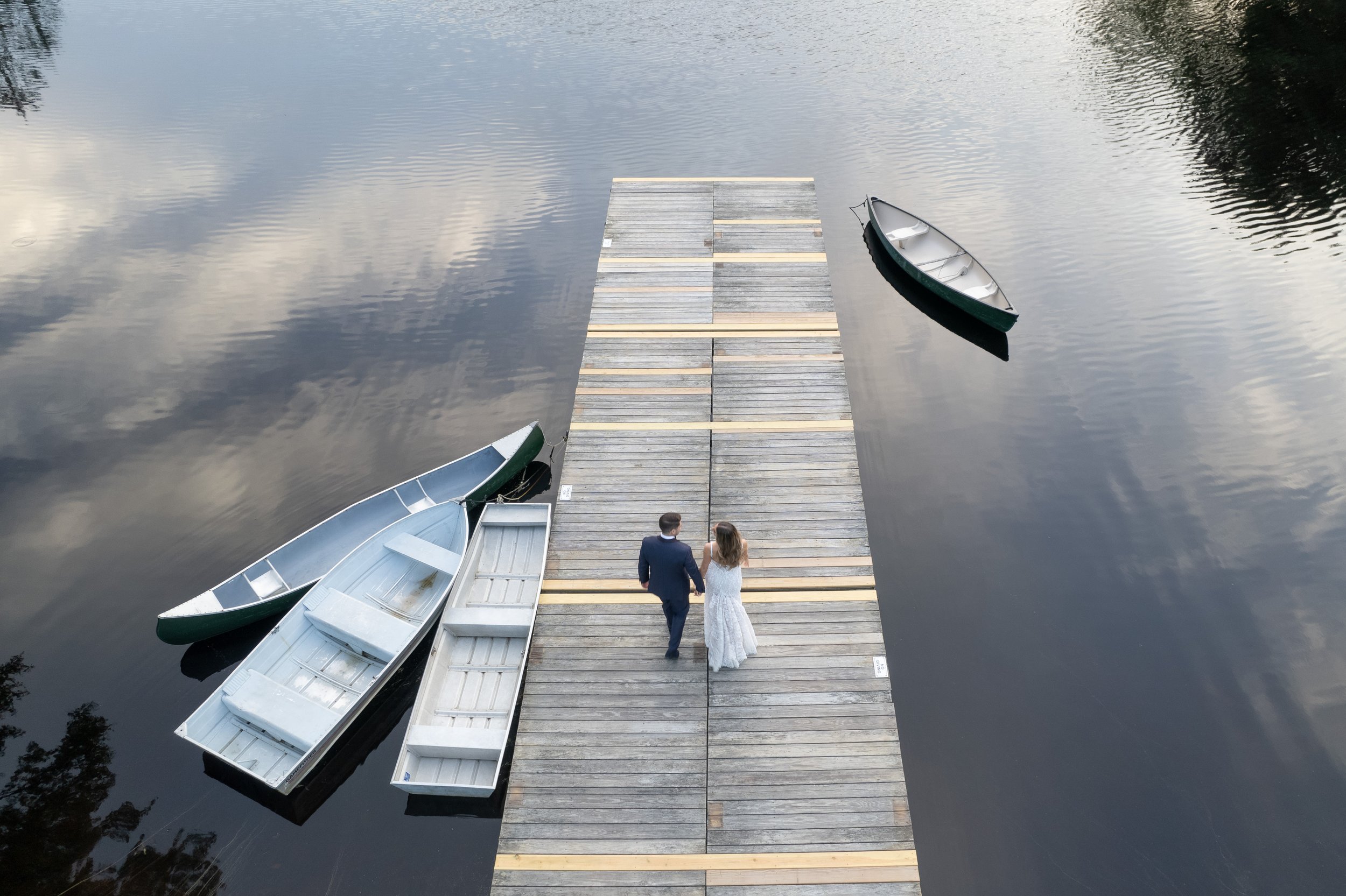 A couple in wedding attire walking hand in hand on a wooden dock over calm water, with two boats tied to the dock and one boat floating nearby.