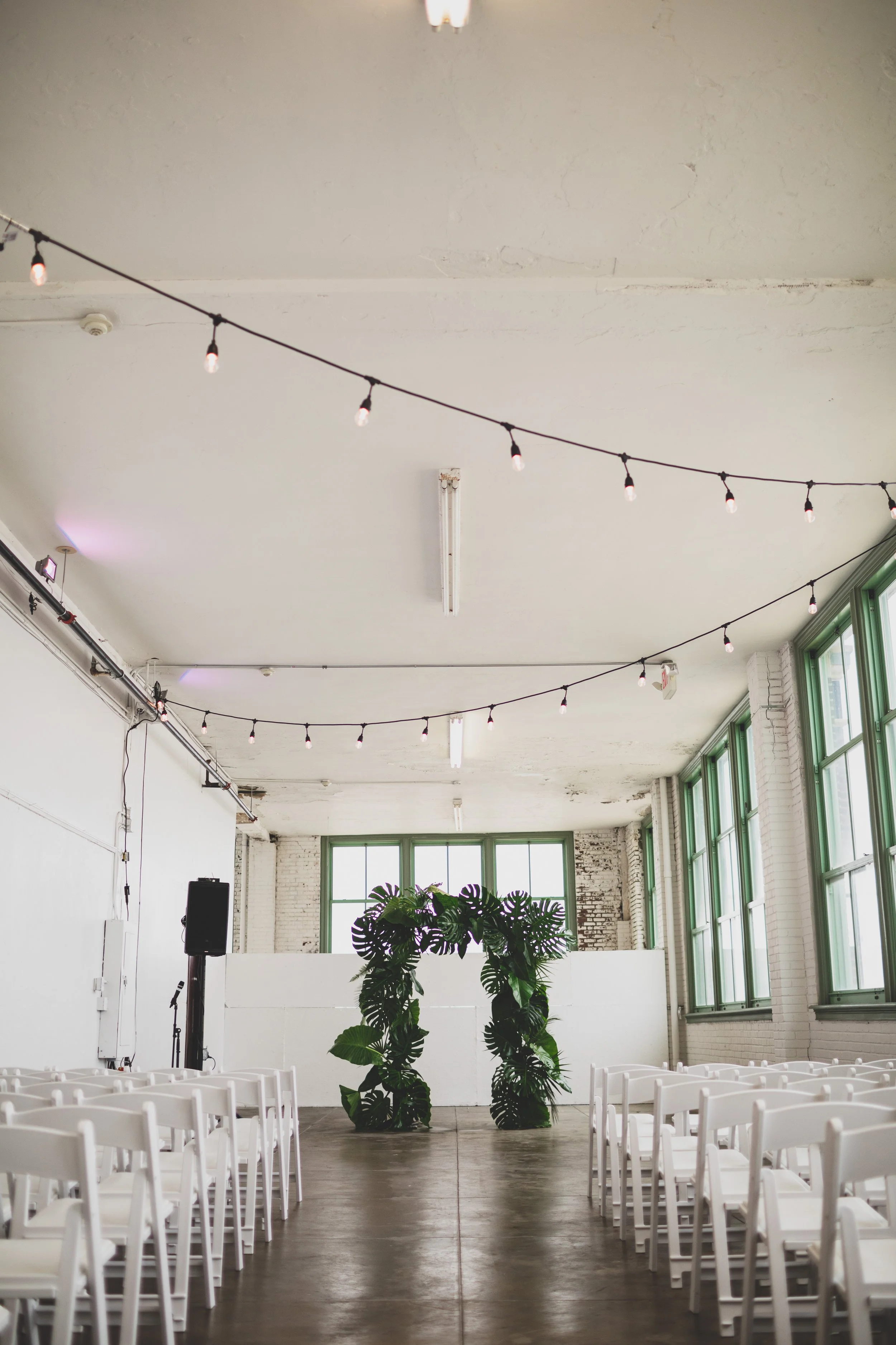 Indoor event space with white chairs arranged on either side of an aisle, decorated with green tropical leaves. String lights hang from the ceiling, and large windows on the right provide natural light.