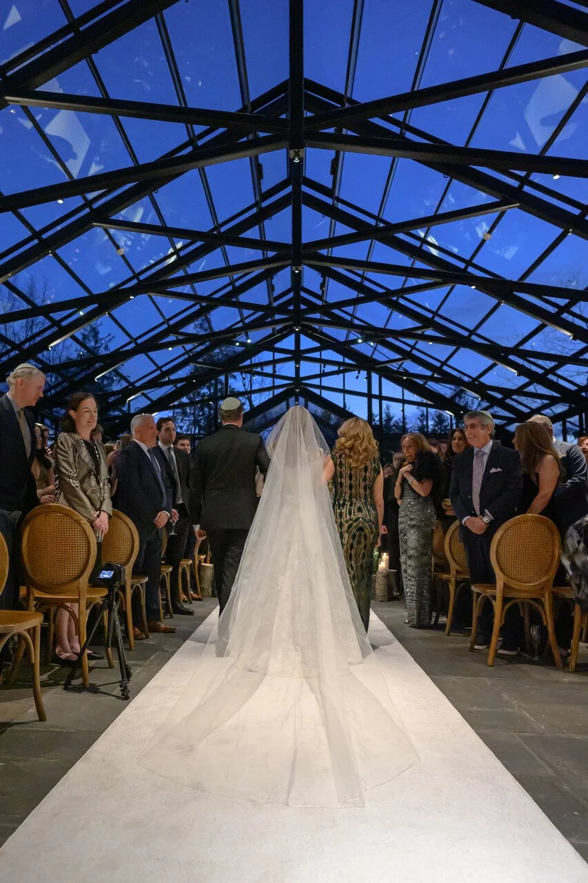 A bride in a wedding gown with a long veil walking down the aisle at her wedding ceremony, with guests standing on either side under a glass ceiling at dusk.