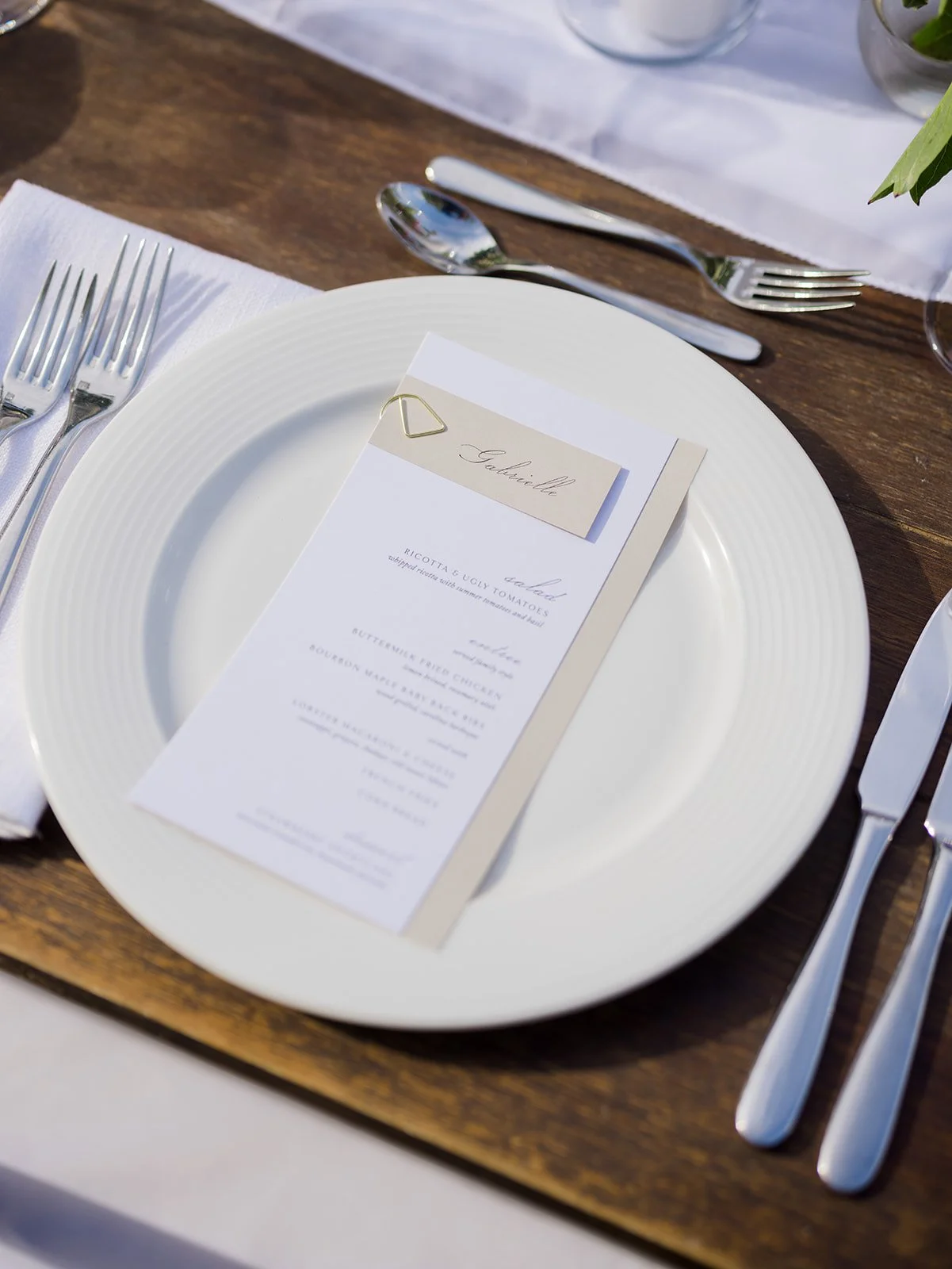 Elegant table setting with a white dinner plate, a printed menu, silverware, and a name card clipped with a gold paperclip, on a wooden table with a white tablecloth.