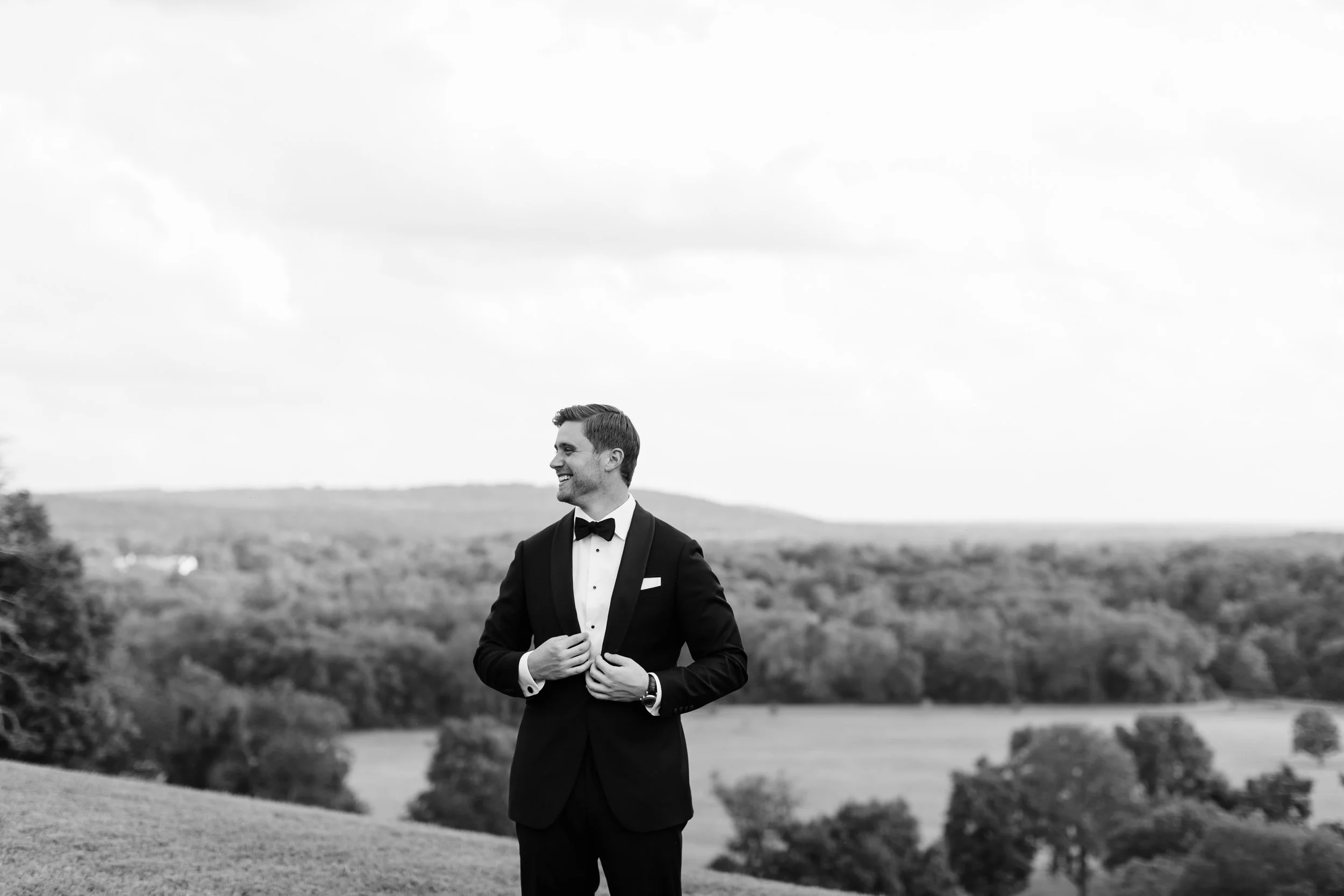 A man in a tuxedo standing outdoors with a scenic landscape background, smiling, adjusting his jacket.