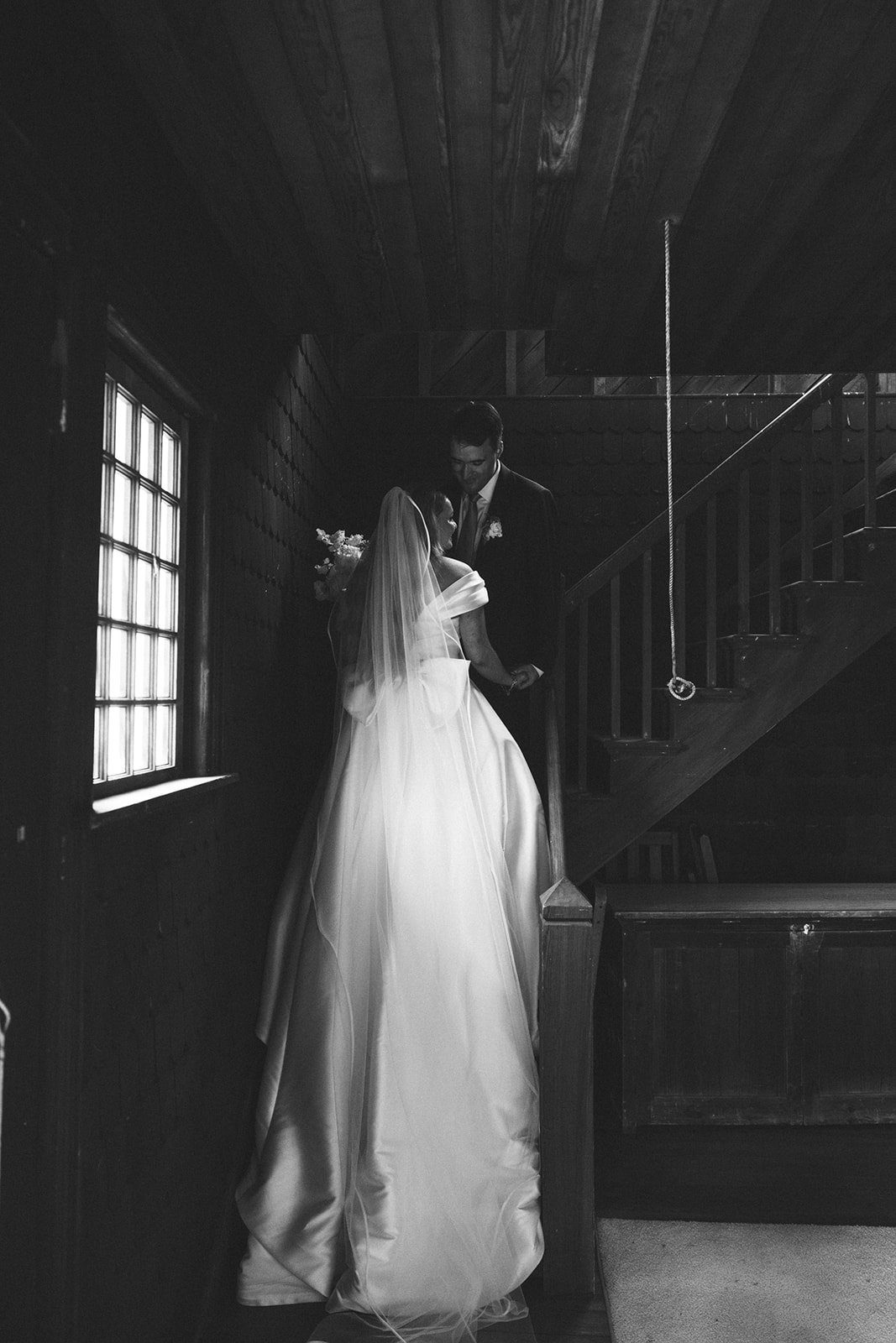 Black and white photo of a bride and groom standing on a staircase inside a wooden building. The bride wears a strapless wedding gown with a veil, and the groom is in a dark suit. They are holding hands, looking at each other.