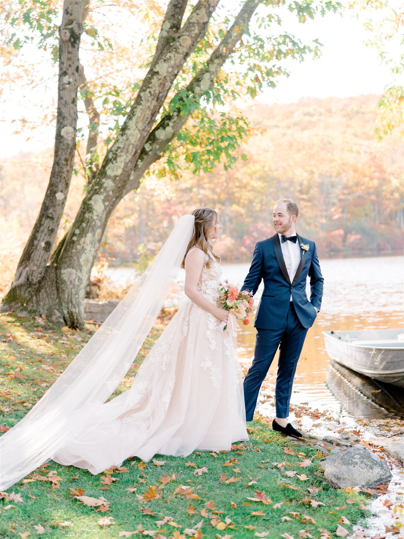A bride and groom standing by a lake surrounded by autumn trees, with the bride holding a bouquet of flowers.