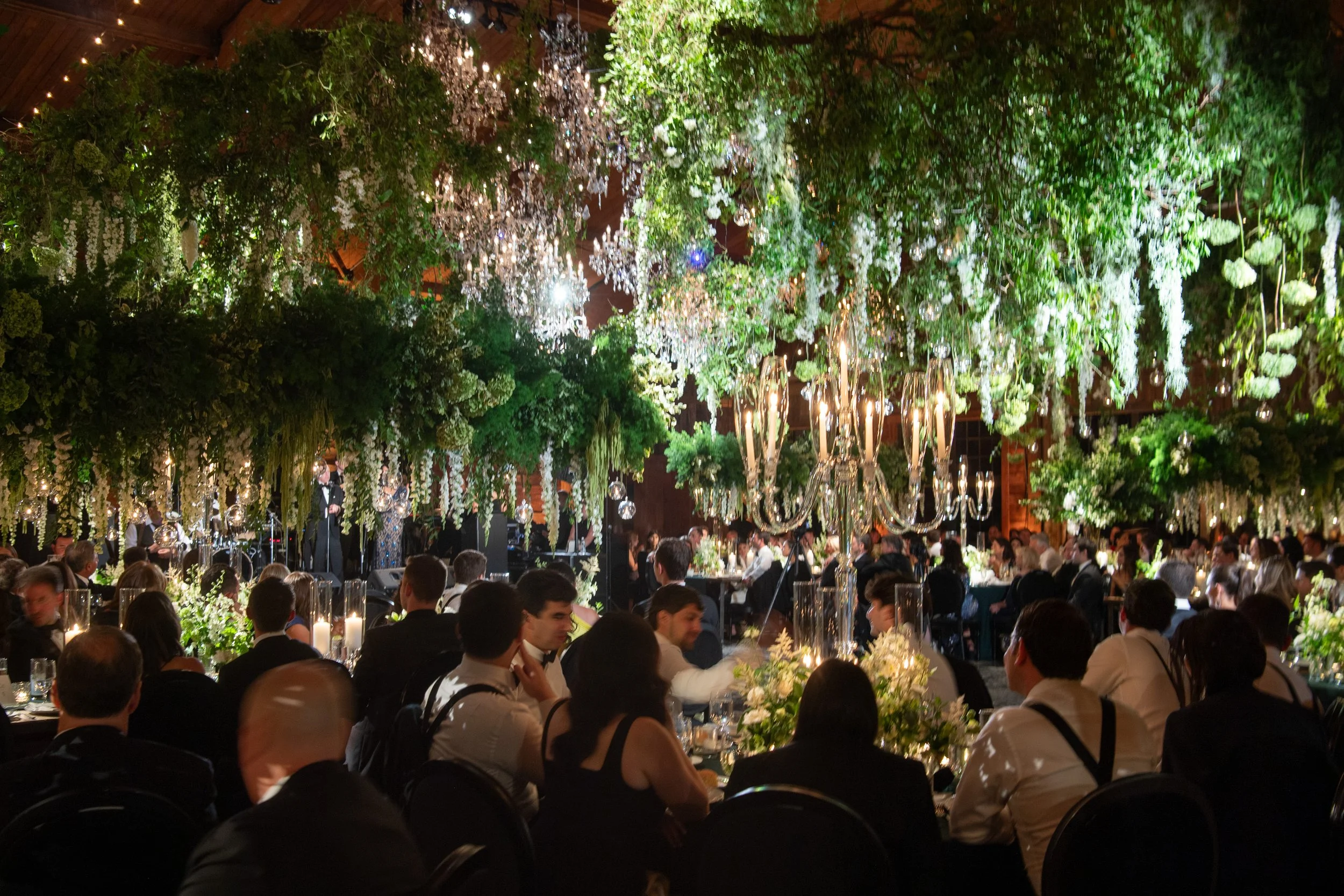 An elegant banquet hall decorated with lush greenery, white flowers, and crystal chandeliers, filled with seated guests attending a formal event or celebration.