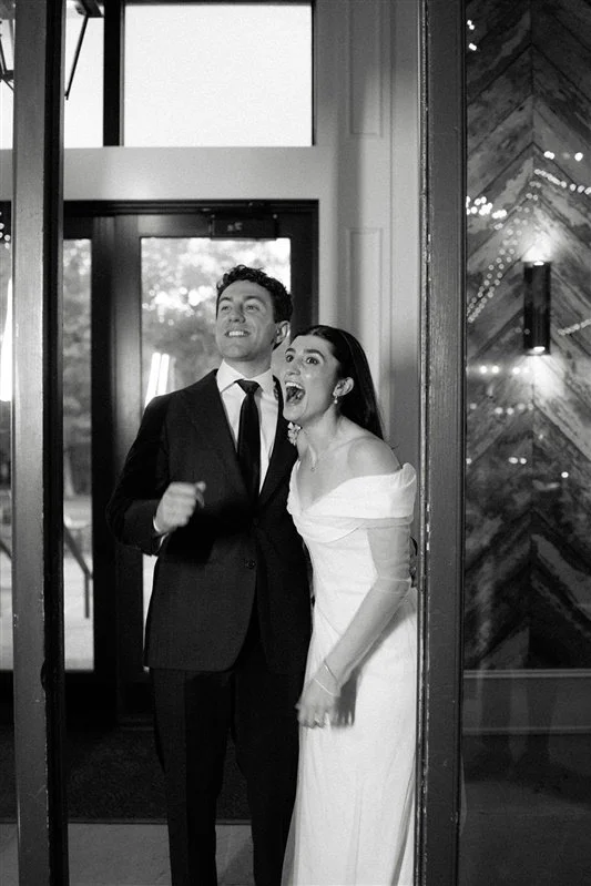 A couple in formal attire, a man in a suit and tie and a woman in a wedding dress, standing at an entrance with a joyful expression.