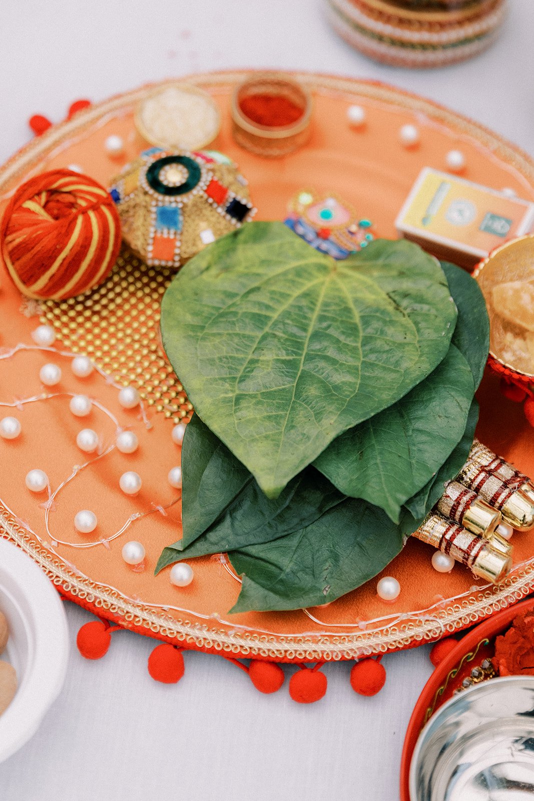 Decorative orange tray with green leaves, pearl string, colorful fabric balls, small gold and silver tubes, a small stack of cash, and other small decorative items.