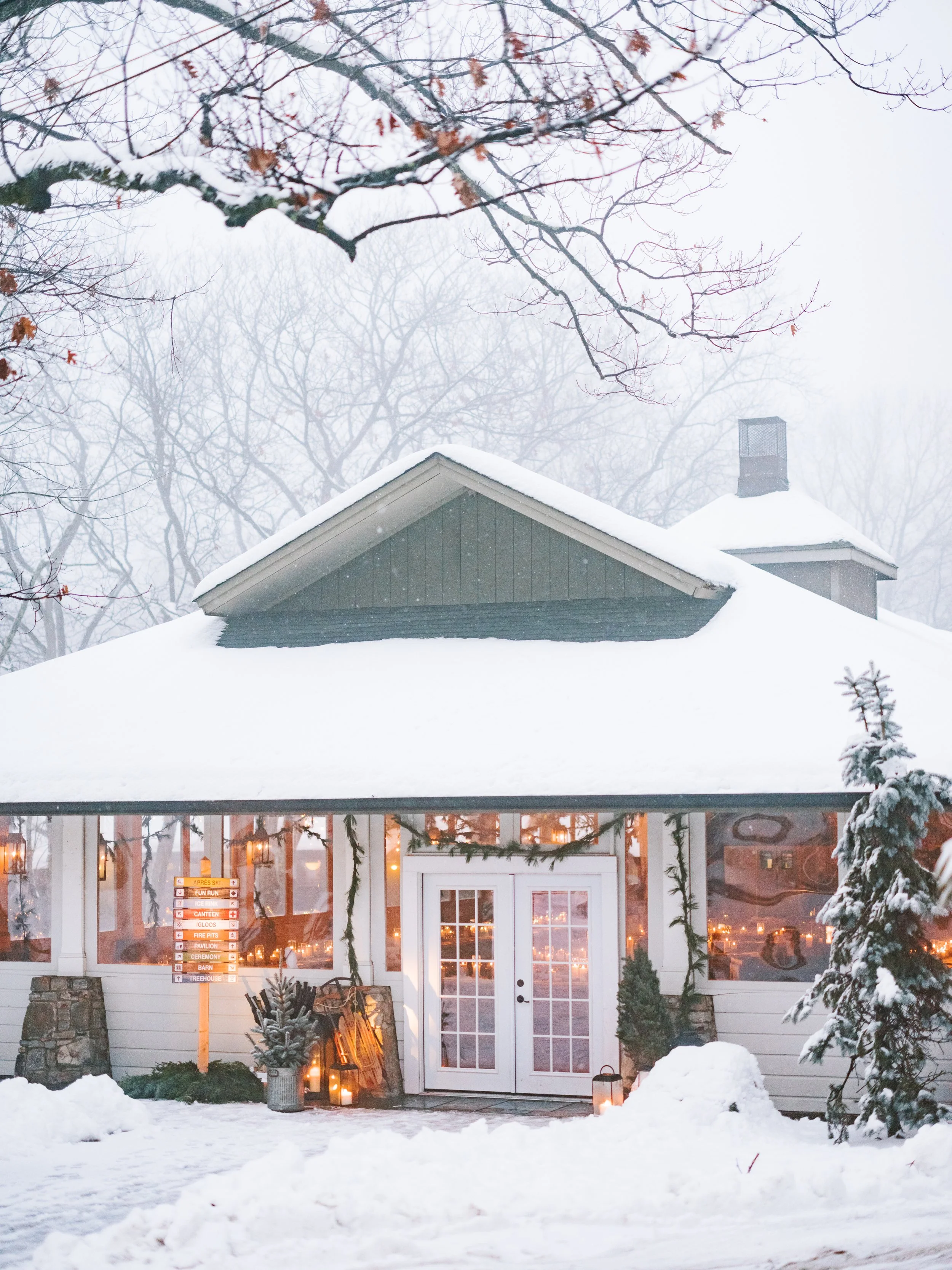 Snow-covered building with illuminated windows, decorated with holiday greenery and candles outside, breezy winter scene with bare trees and falling snow.