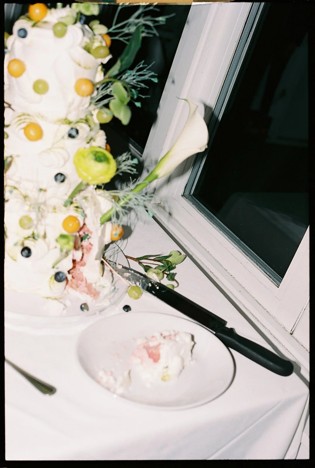 Partially eaten cake with white frosting, decorated with green and yellow berries, on a white table near a window with white frame and a white lily flower.