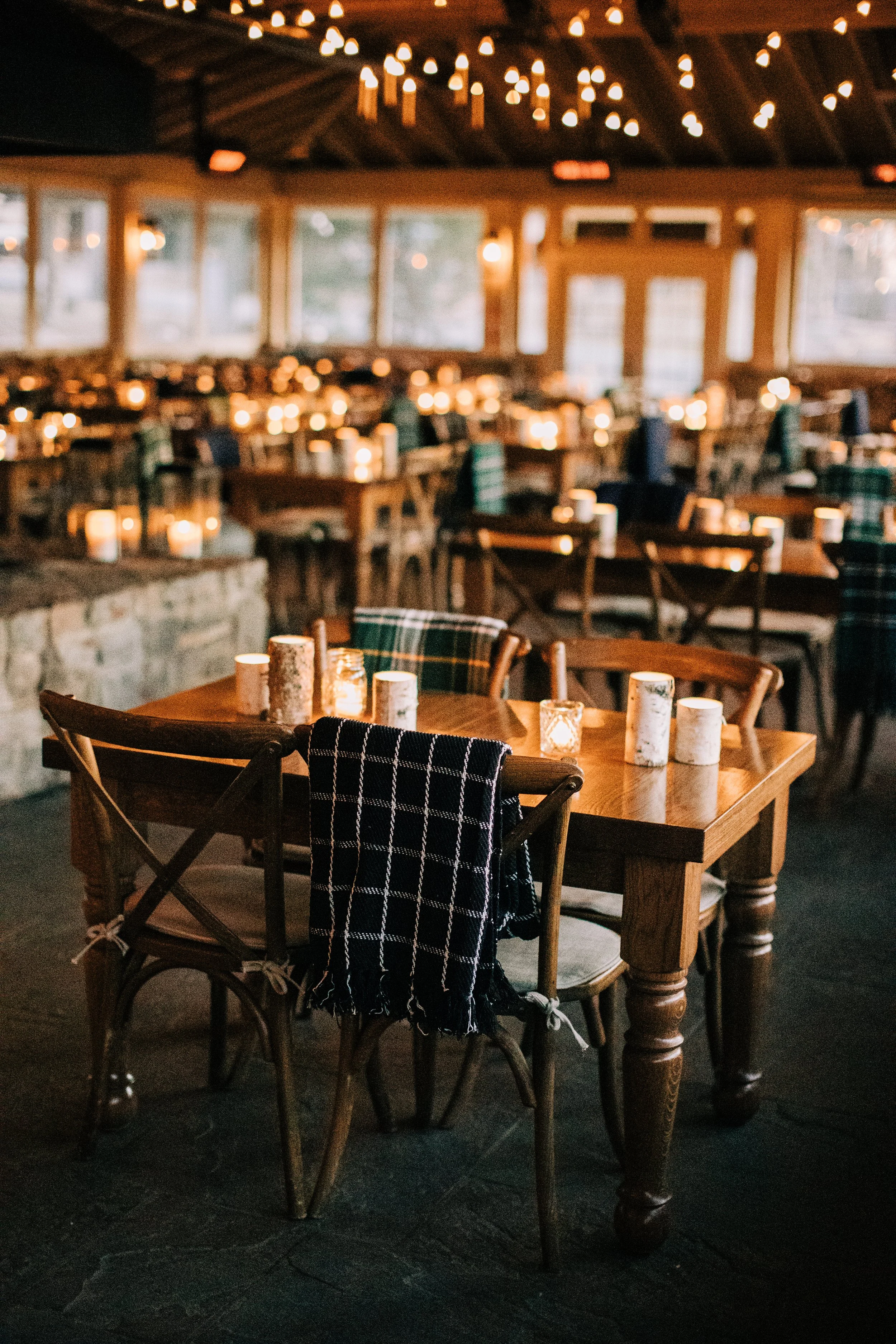 A cozy restaurant with wooden tables and chairs, lit by candles and string lights, with large windows in the background.