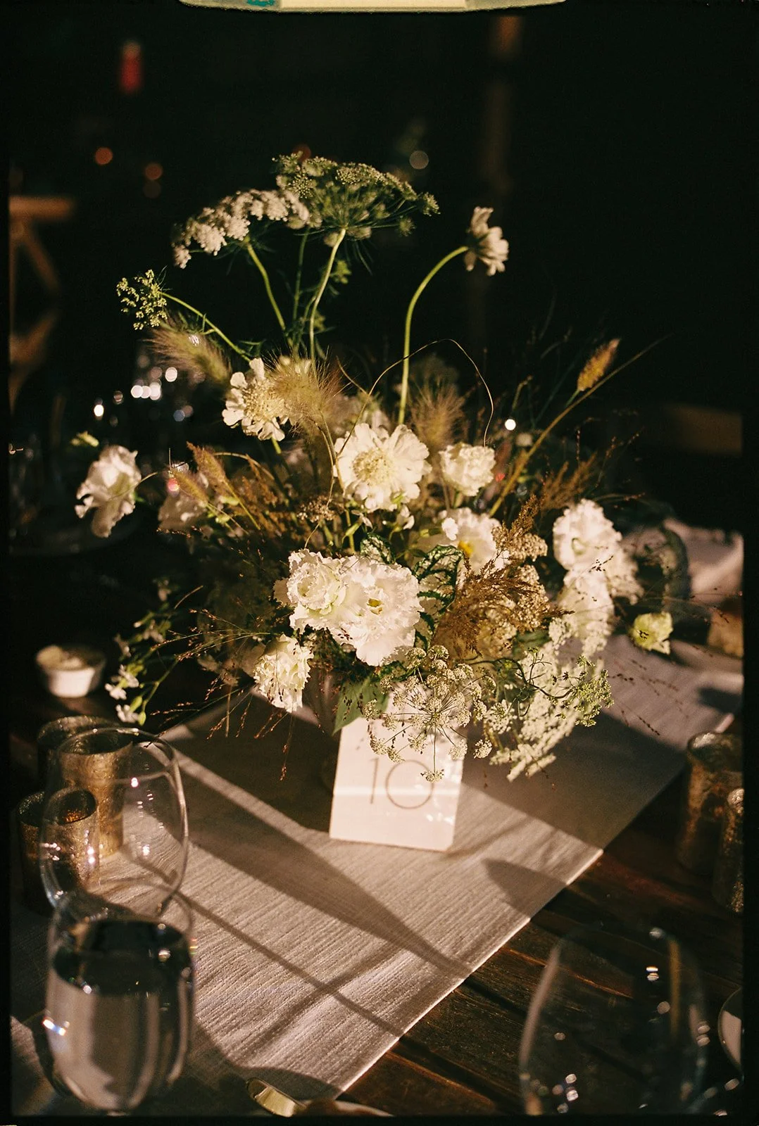 A floral centerpiece with white flowers and greenery on a dark-colored table, illuminated by warm lighting at a nighttime event.