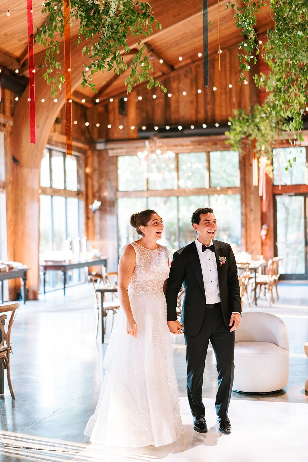 A bride and groom holding hands and smiling inside a rustic wooden wedding venue decorated with greenery and hanging ribbons.