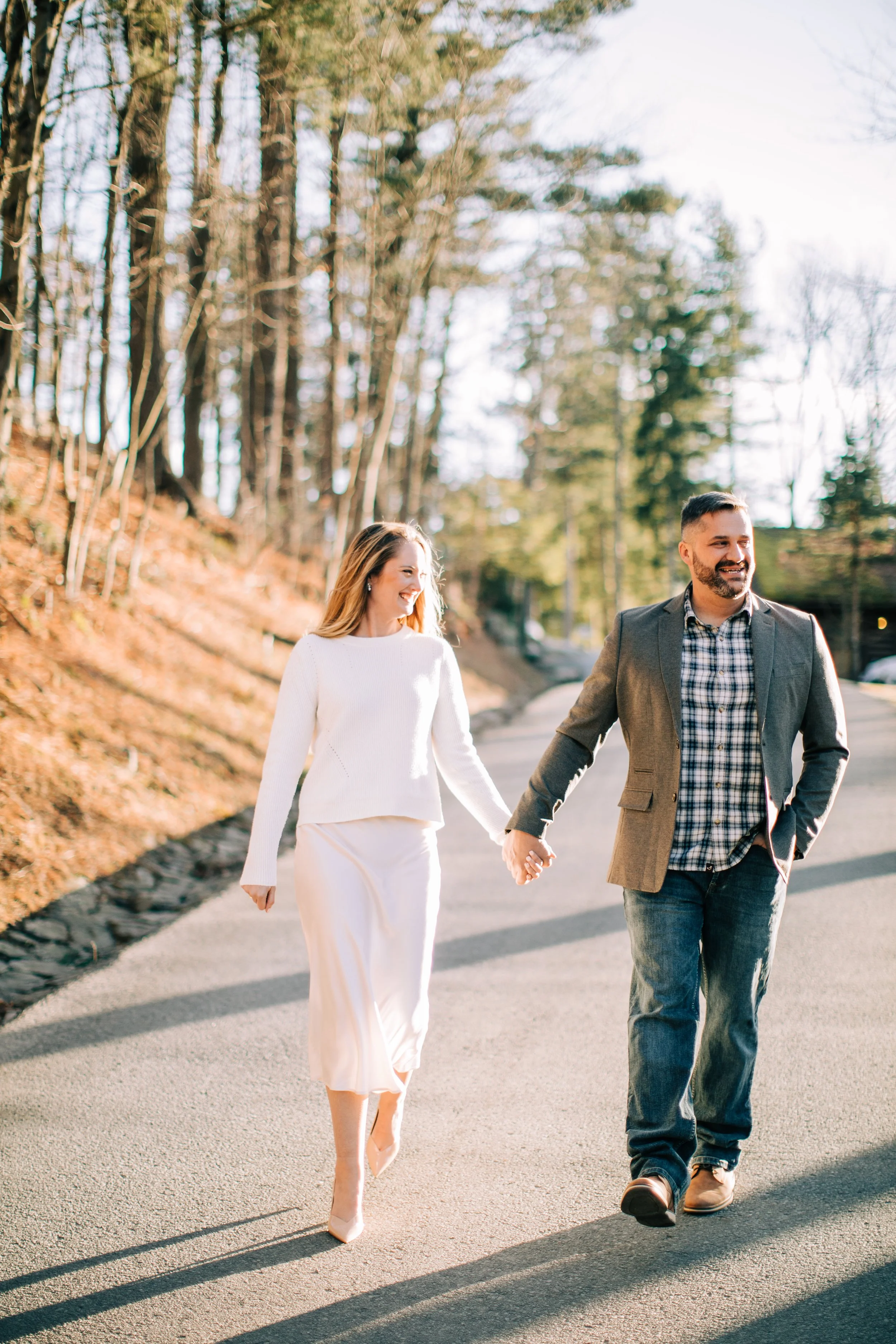 A couple holding hands and walking outdoors on a paved path, surrounded by trees, smiling and enjoying a sunny day.