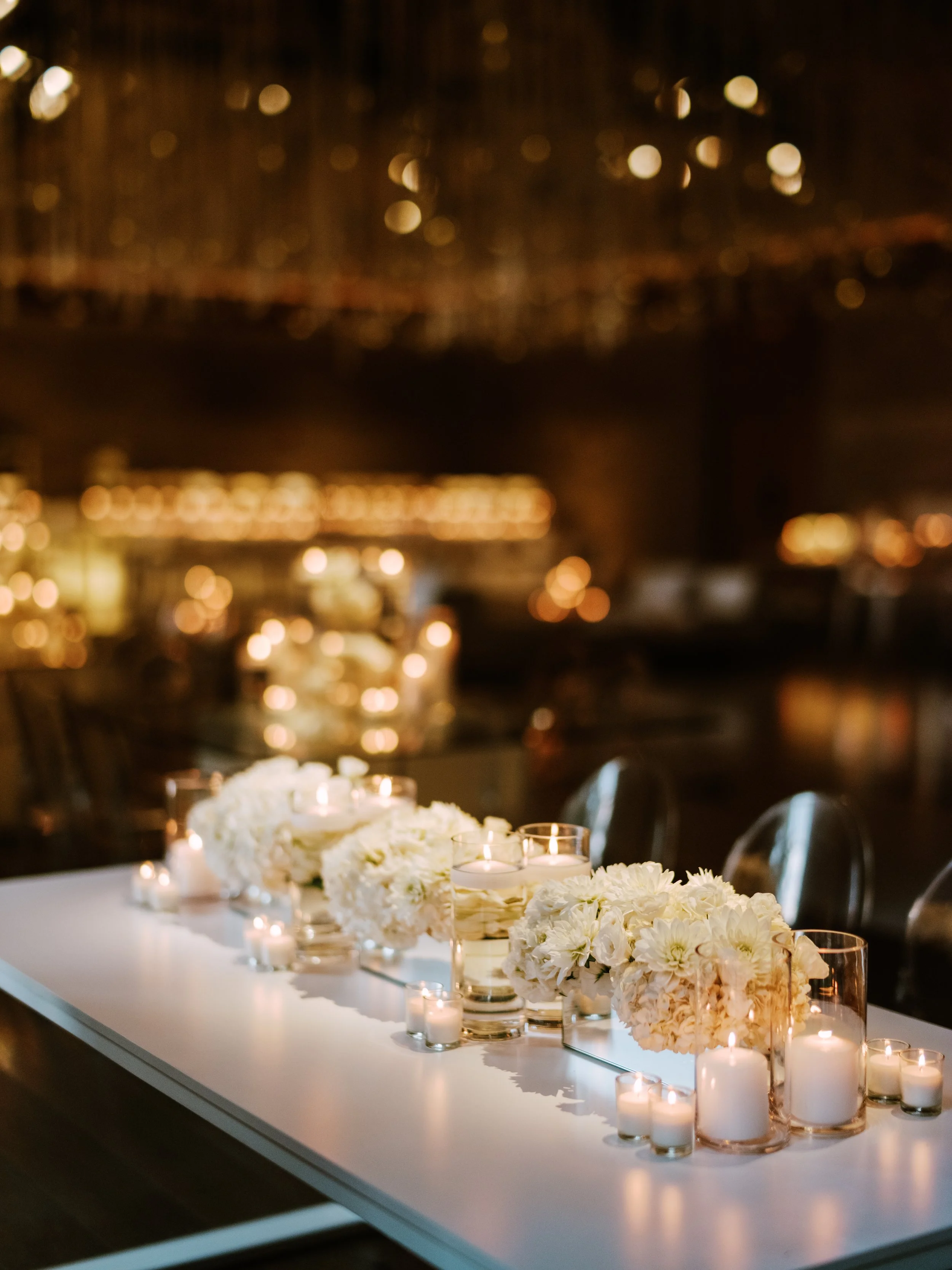 Table decorated with white flowers and candles in glass holders at an elegant event, with a blurred background of warm lights.