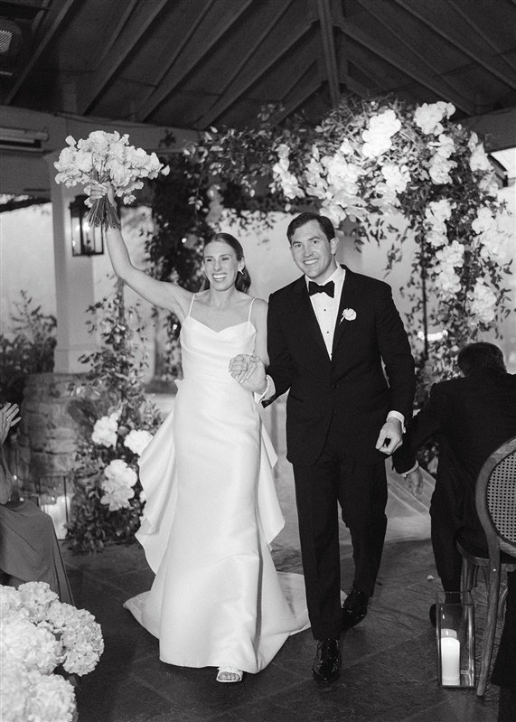 Black and white photo of a bride and groom walking arm in arm at their wedding reception, with the bride holding a bouquet of flowers and smiling, while the groom is dressed in a tuxedo and smiling. They are under a floral arch and guests are seated 