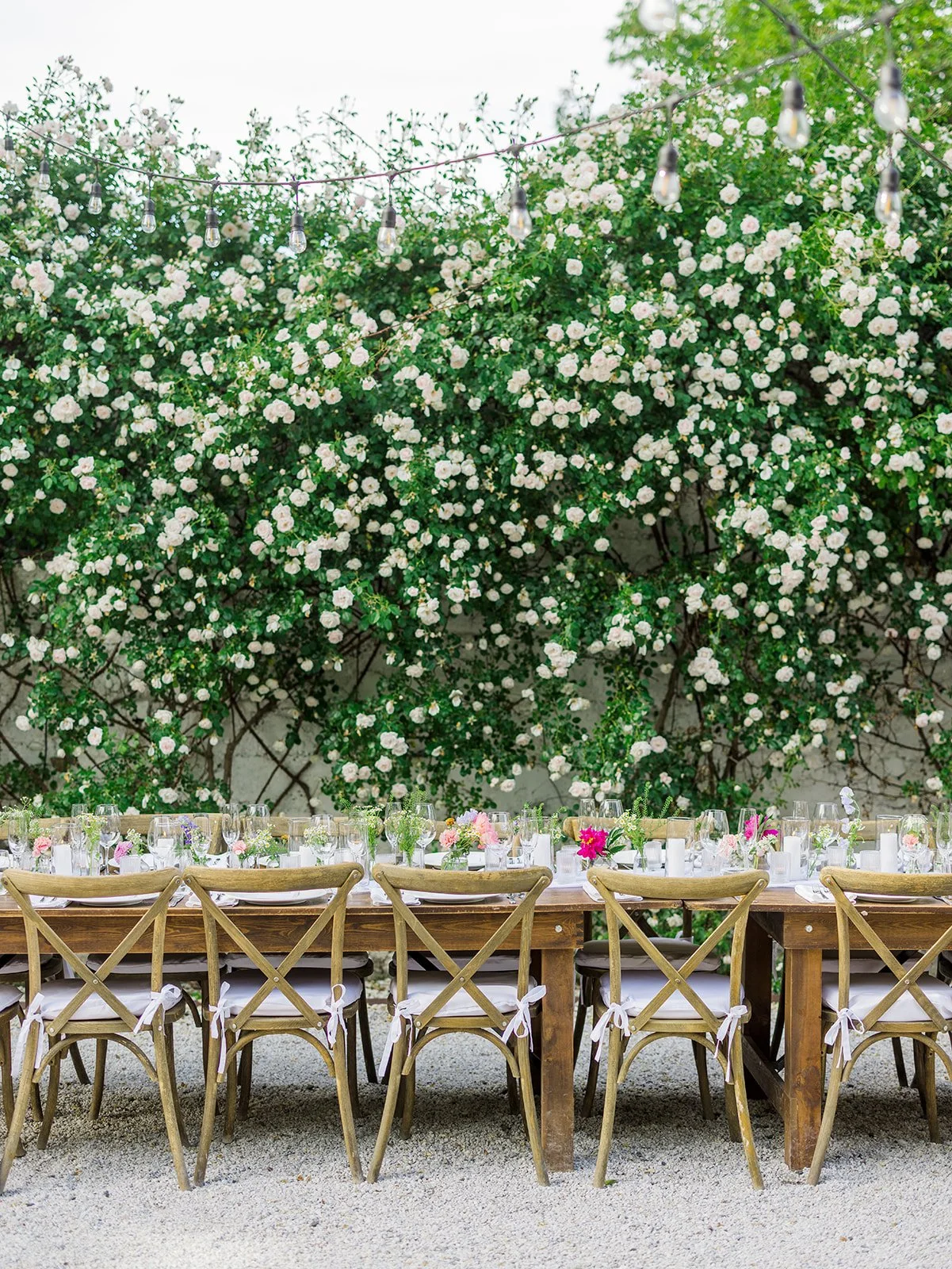 Outdoor dining setup with a long wooden table and chairs, decorated with flowers, against a backdrop of lush green foliage and white blooming flowers, with string lights overhead.