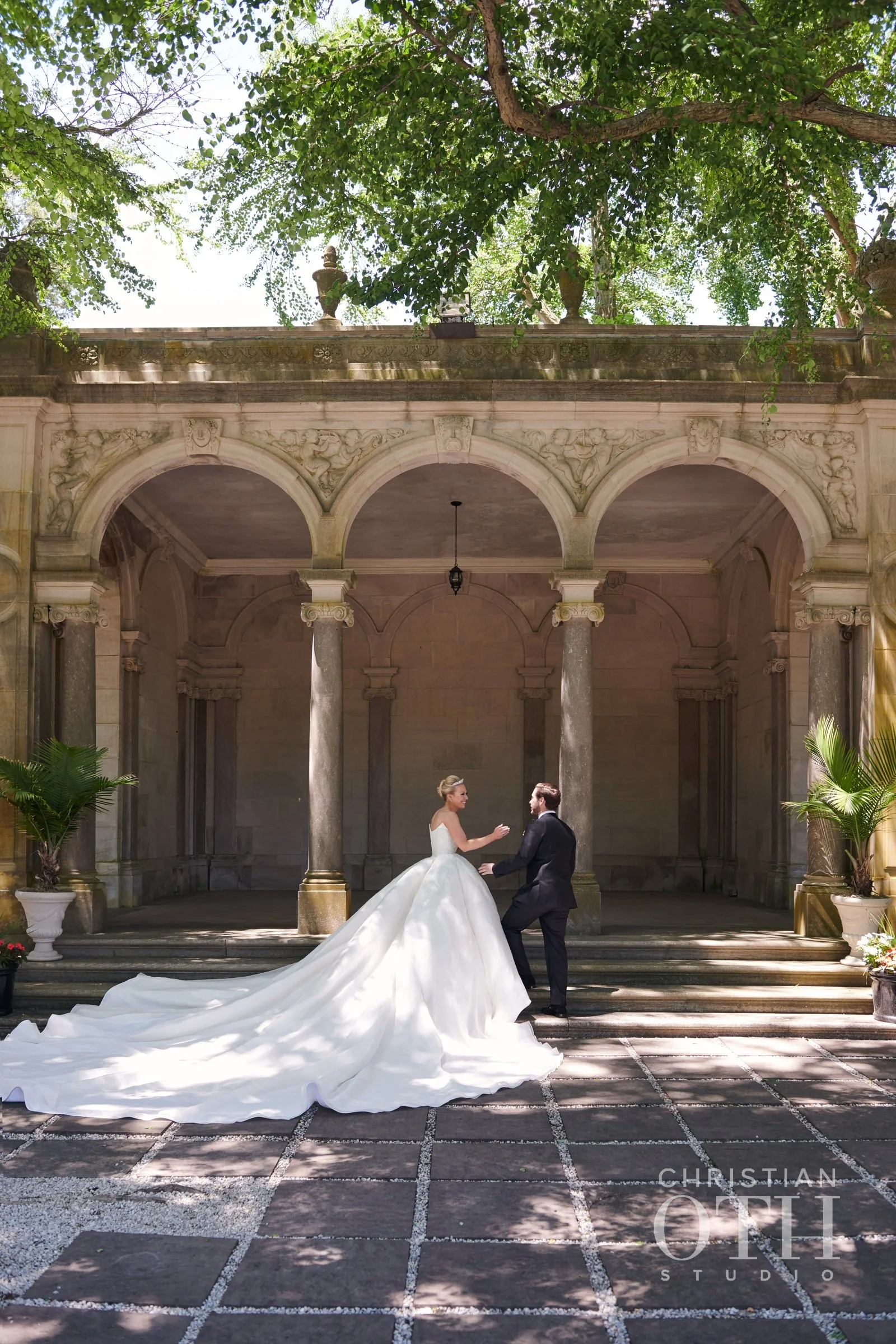 A bride in a white wedding gown with a long train and a groom in a black suit holding hands on the steps of an ornate stone pavilion with columns, surrounded by greenery.