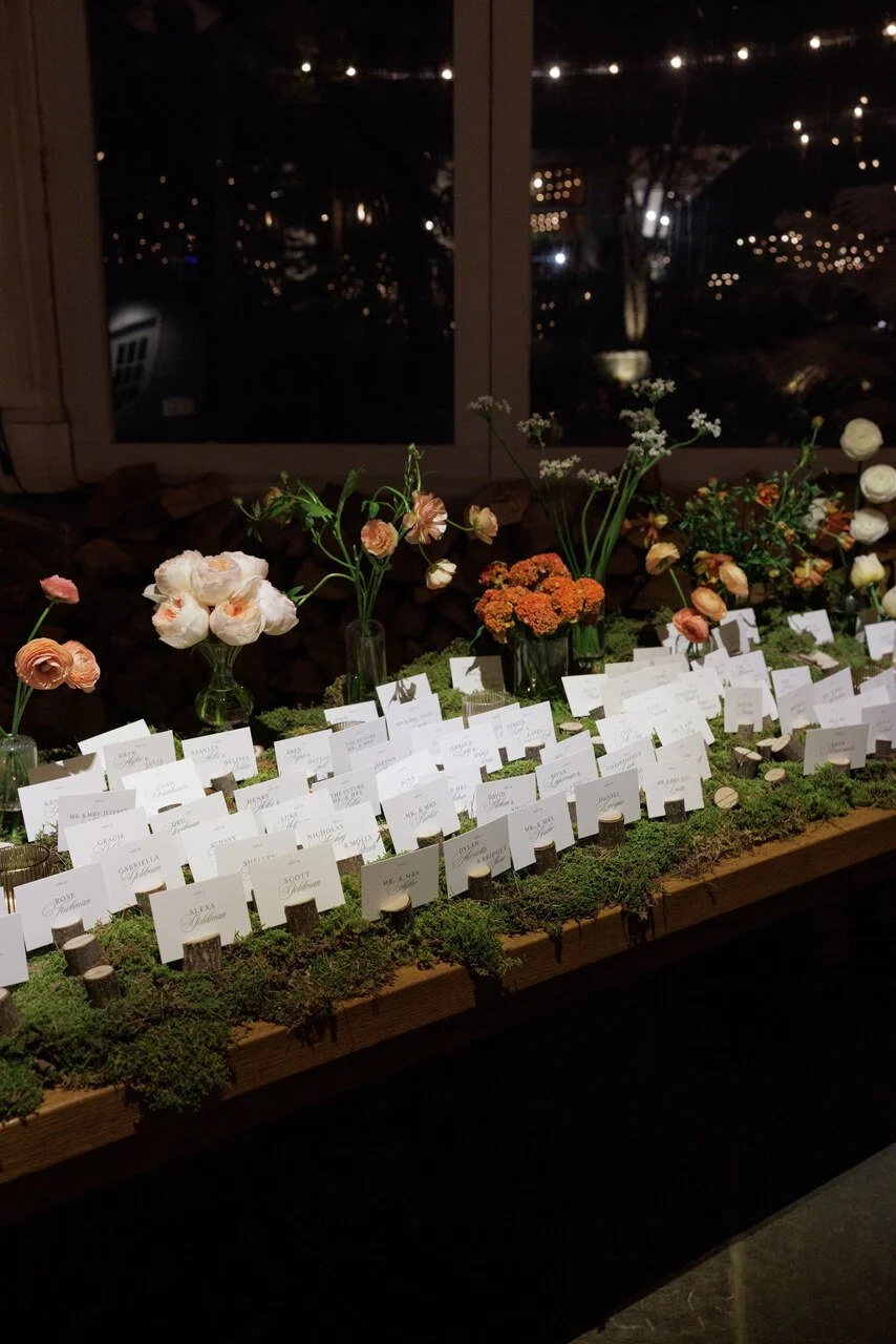 A wedding reception table with floral arrangements in small vases, name cards, and moss-covered surface inside, with a night view through the window showing string lights outside.