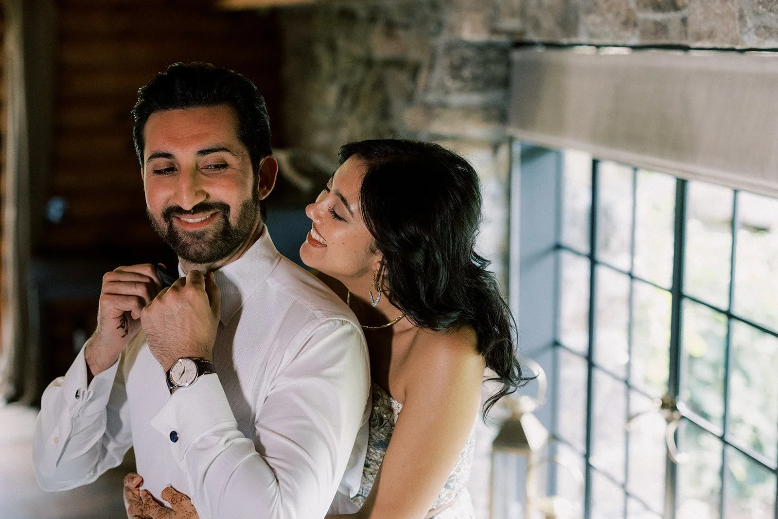 A man with a beard and a woman with dark hair, smiling and close together, indoors near a large window, celebrating.