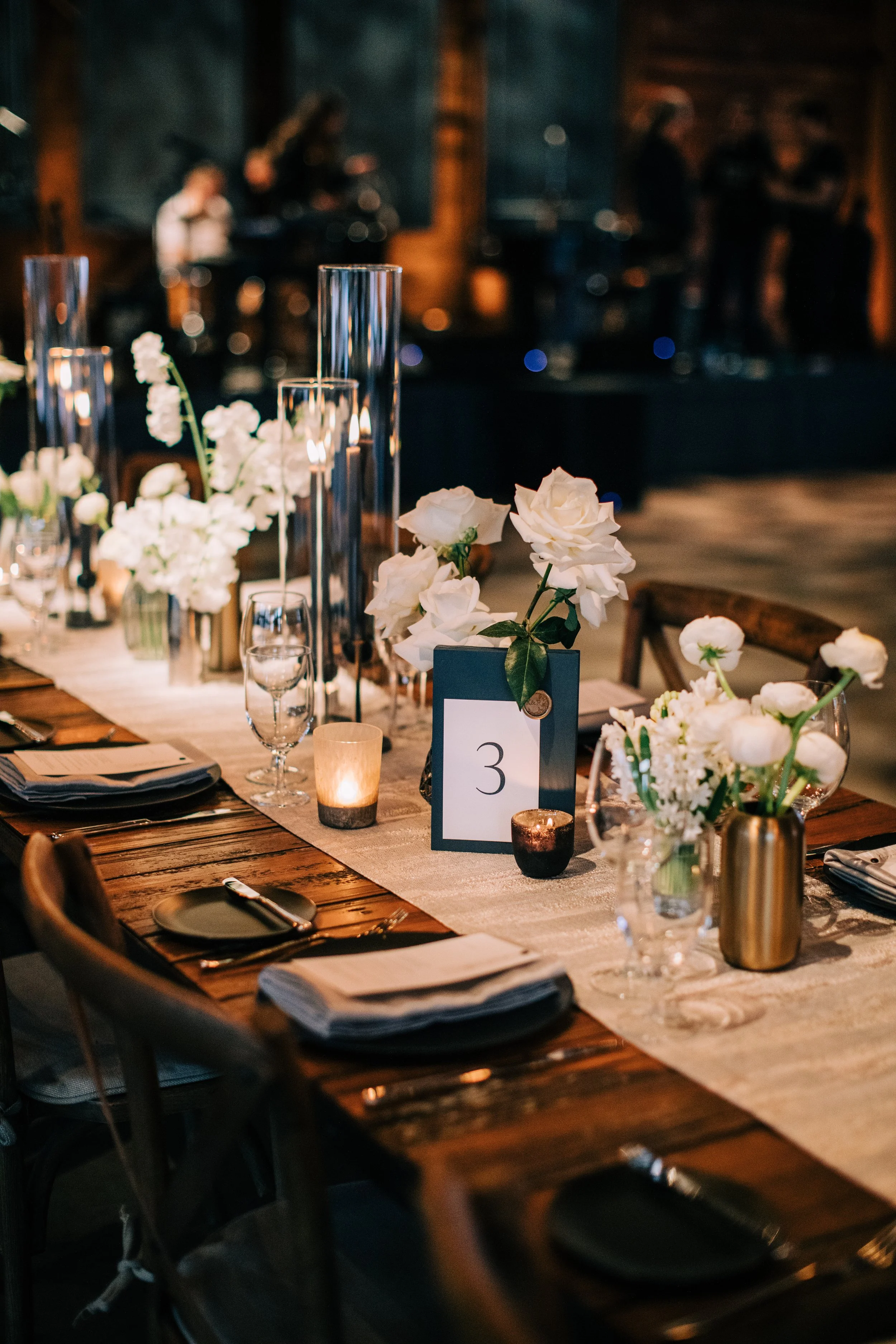 A decorated table at a formal event with white floral centerpieces, candles, wine glasses, black plates, napkins, and table number 3, set in a dimly lit venue with a live band performing in the background.