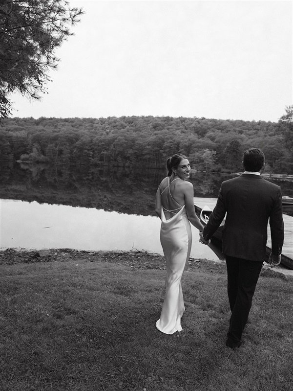 A couple in formal attire, walking hand in hand by a lake at sunset or dusk, with trees in the background.