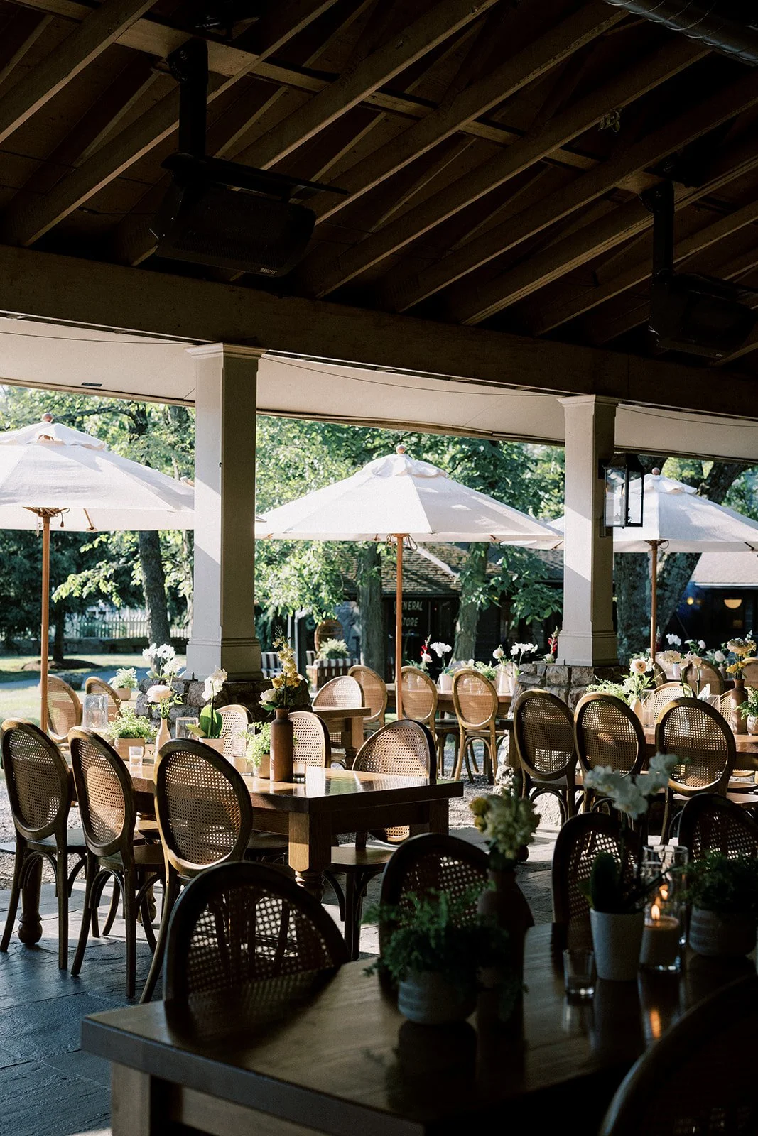 Outdoor patio with tables and chairs, decorated with floral arrangements, under white umbrellas, with trees in the background.