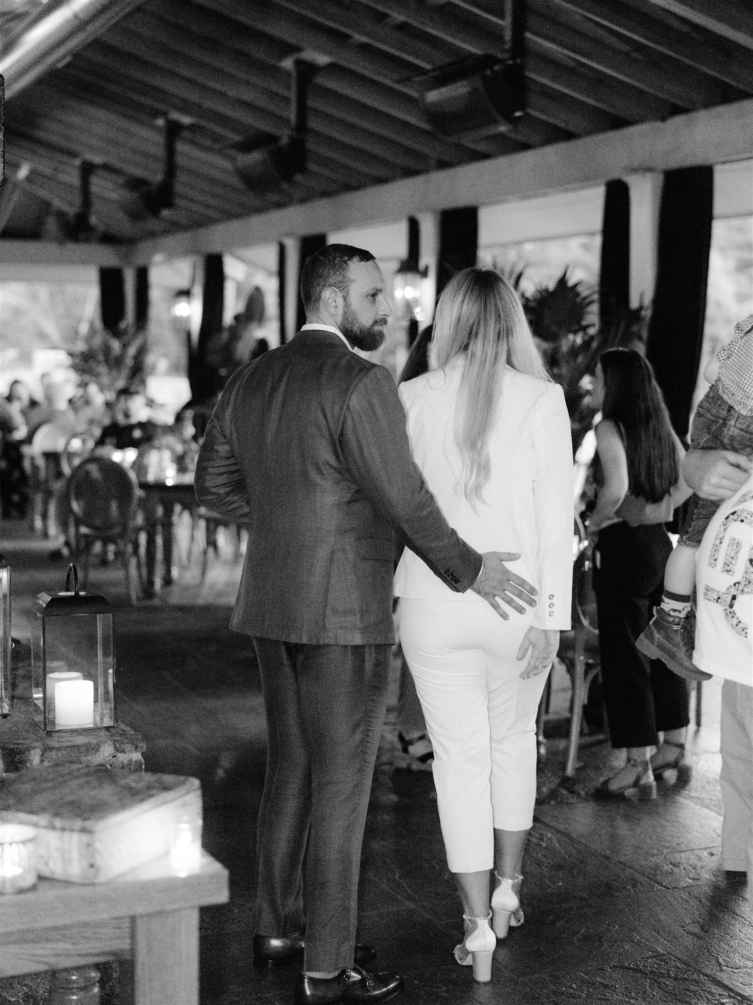 A man in a suit with a beard places his hand on a woman's back as they walk together at a formal event.